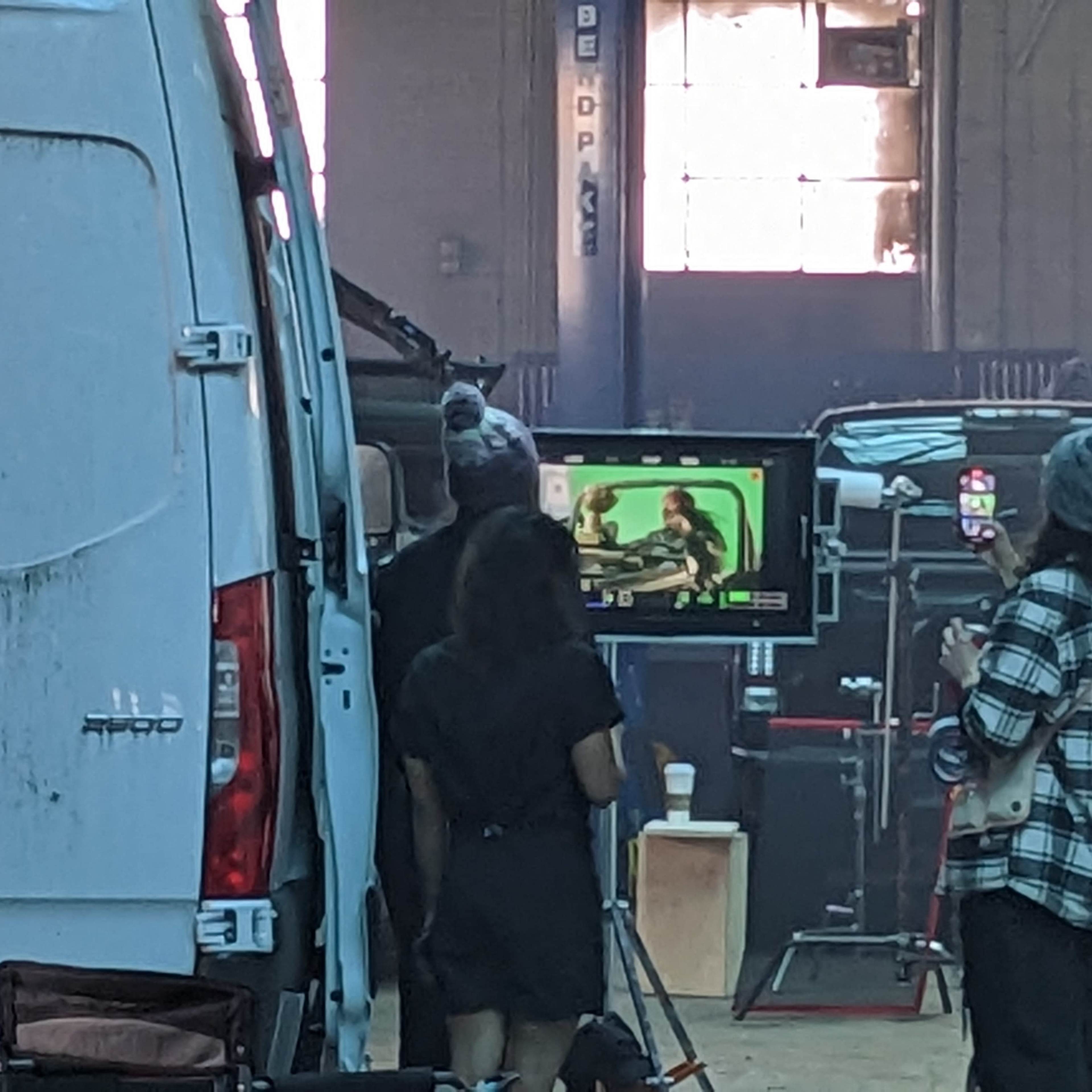 A group of people watches a scene being filmed on a monitor inside a warehouse.