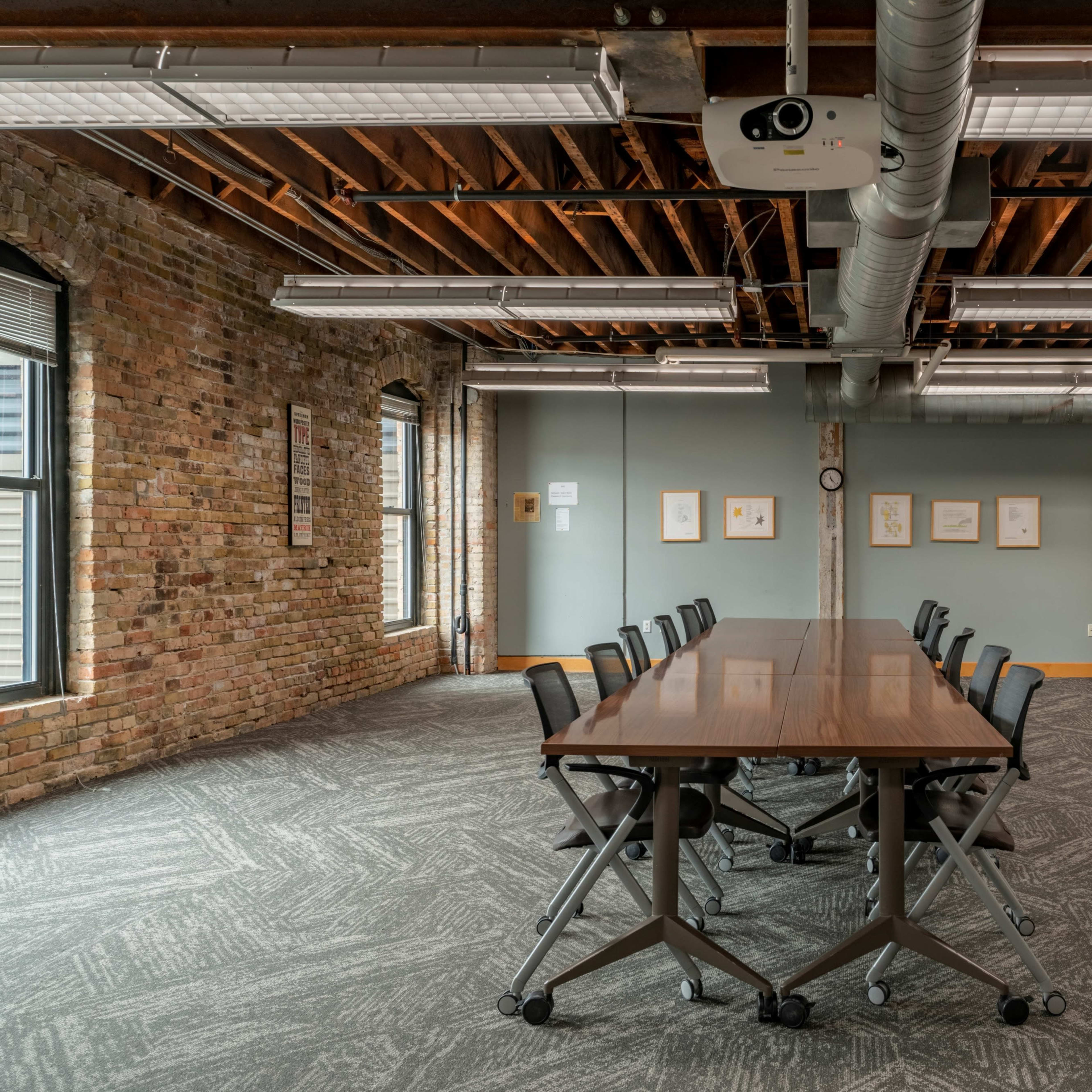 A conference room features a long wooden table surrounded by black chairs, with exposed brick walls and overhead lighting.