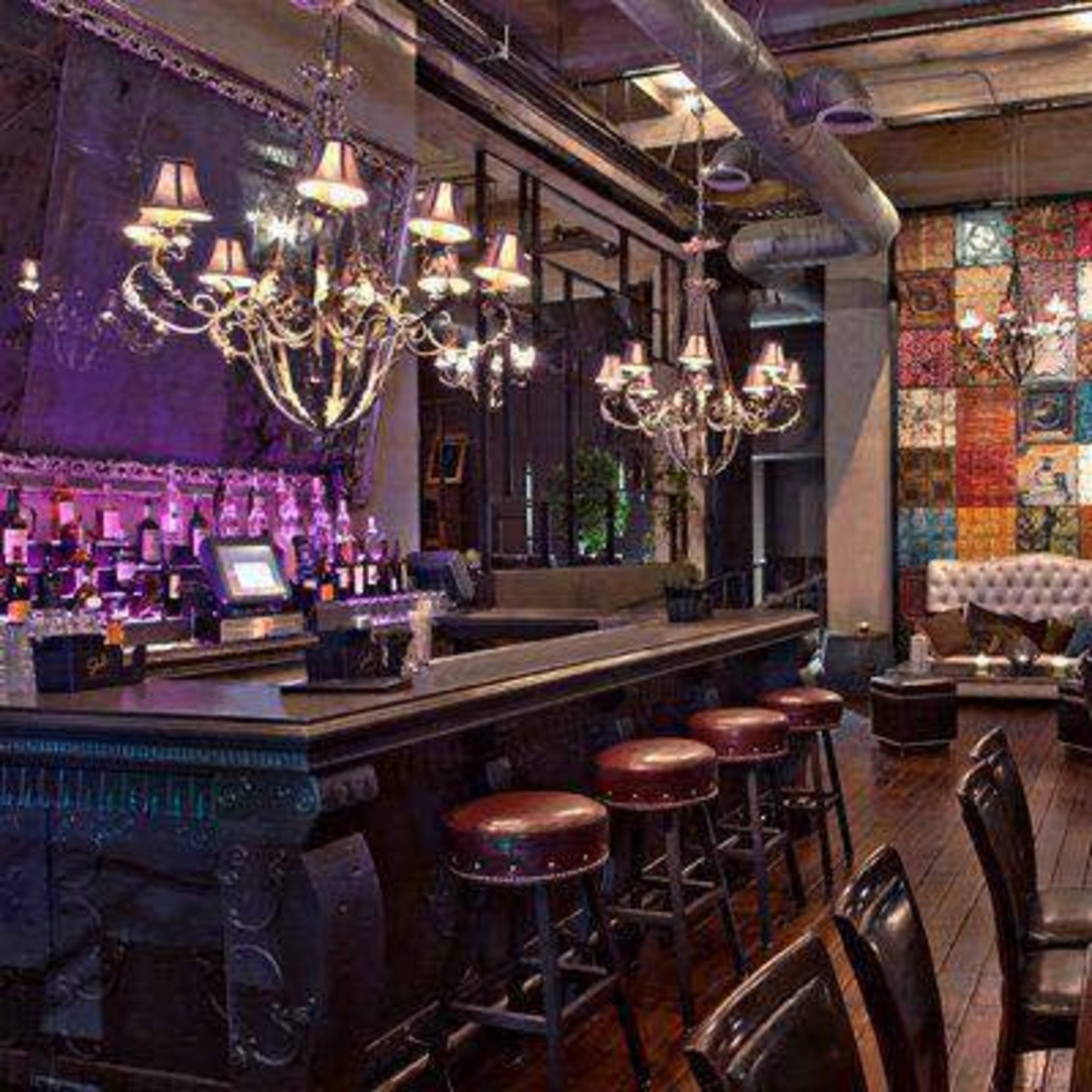 A dimly lit bar with a polished counter, high stools, and decorative chandeliers, complemented by a colorful wall art piece and a lounge area with a tufted sofa.