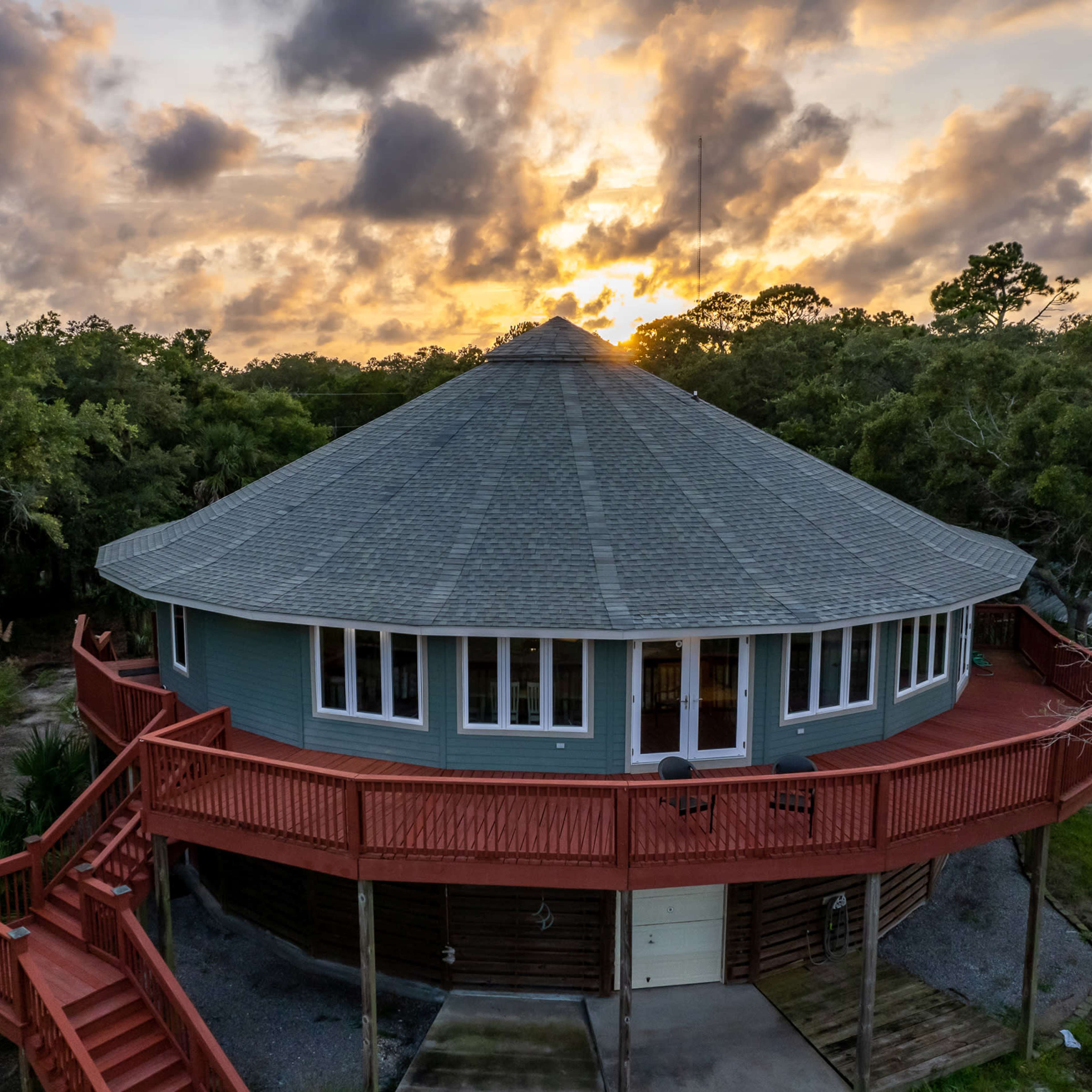 A round, elevated house with a gray roof and a red deck is surrounded by trees at sunset.
