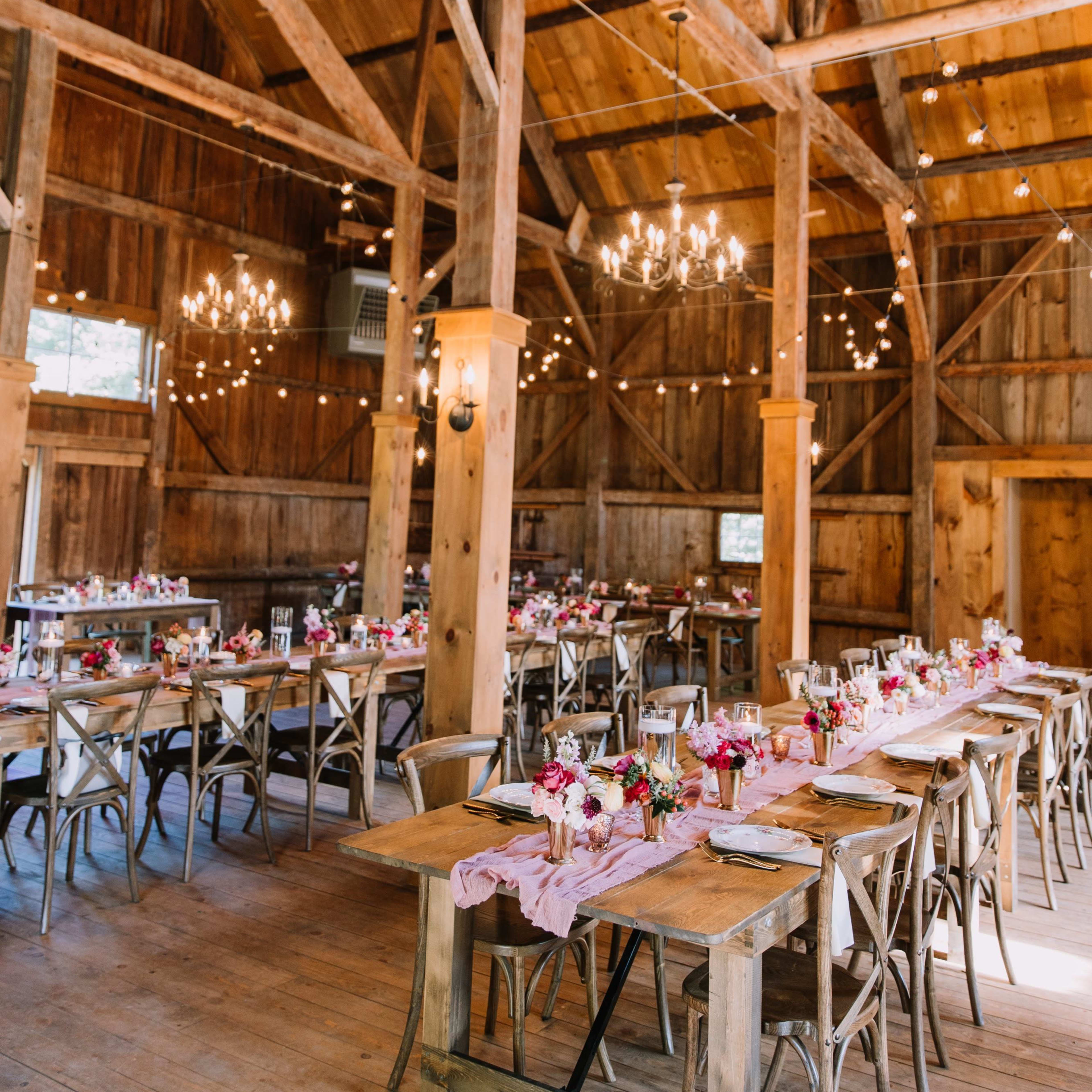 The interior of a rustic barn features long dining tables with floral arrangements, wooden beams, and chandeliers.