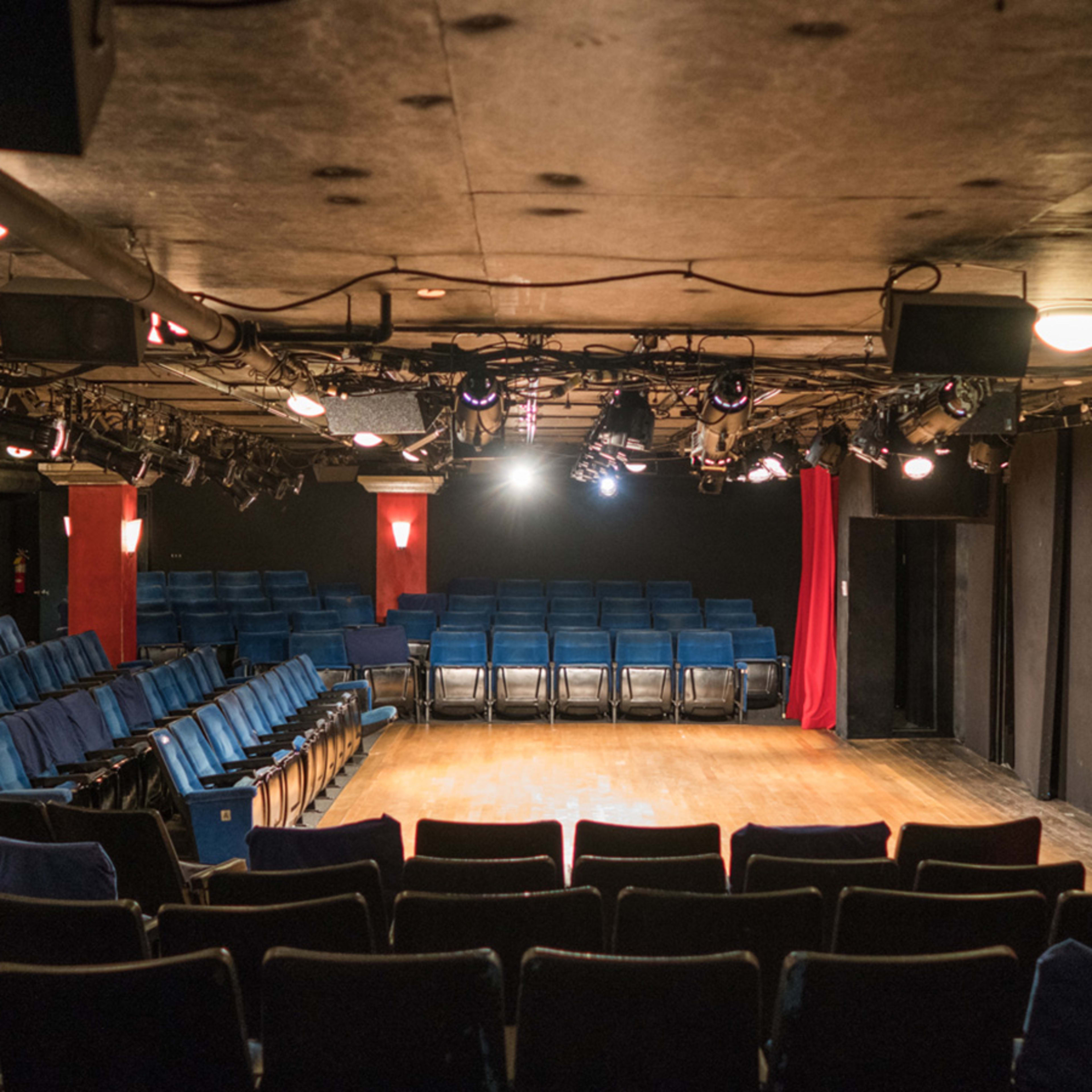 A small theater with rows of blue chairs facing a wooden stage, flanked by red curtains and overhead lighting.