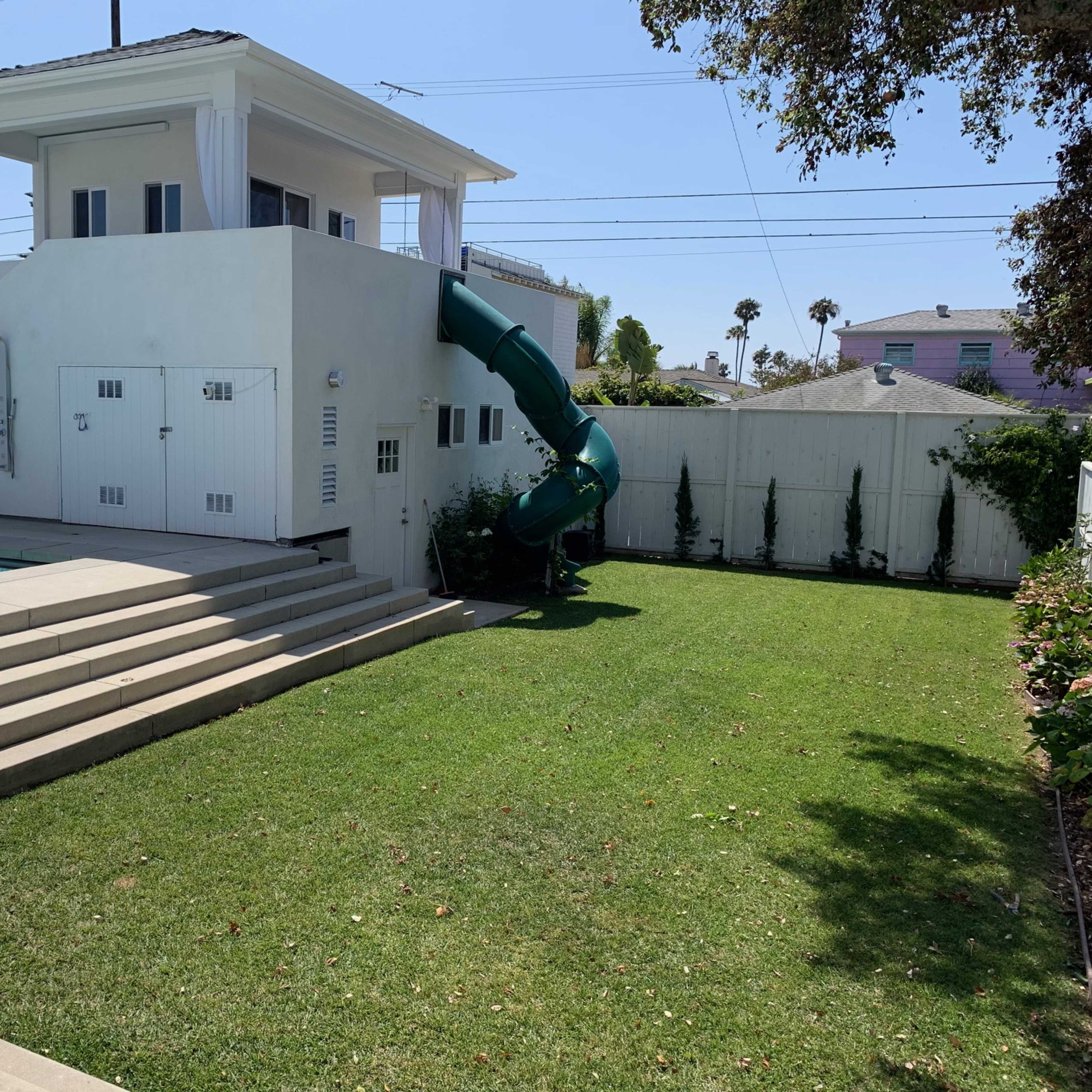 A backyard with a swimming pool, a green waterslide, and a two-story white building in the background.