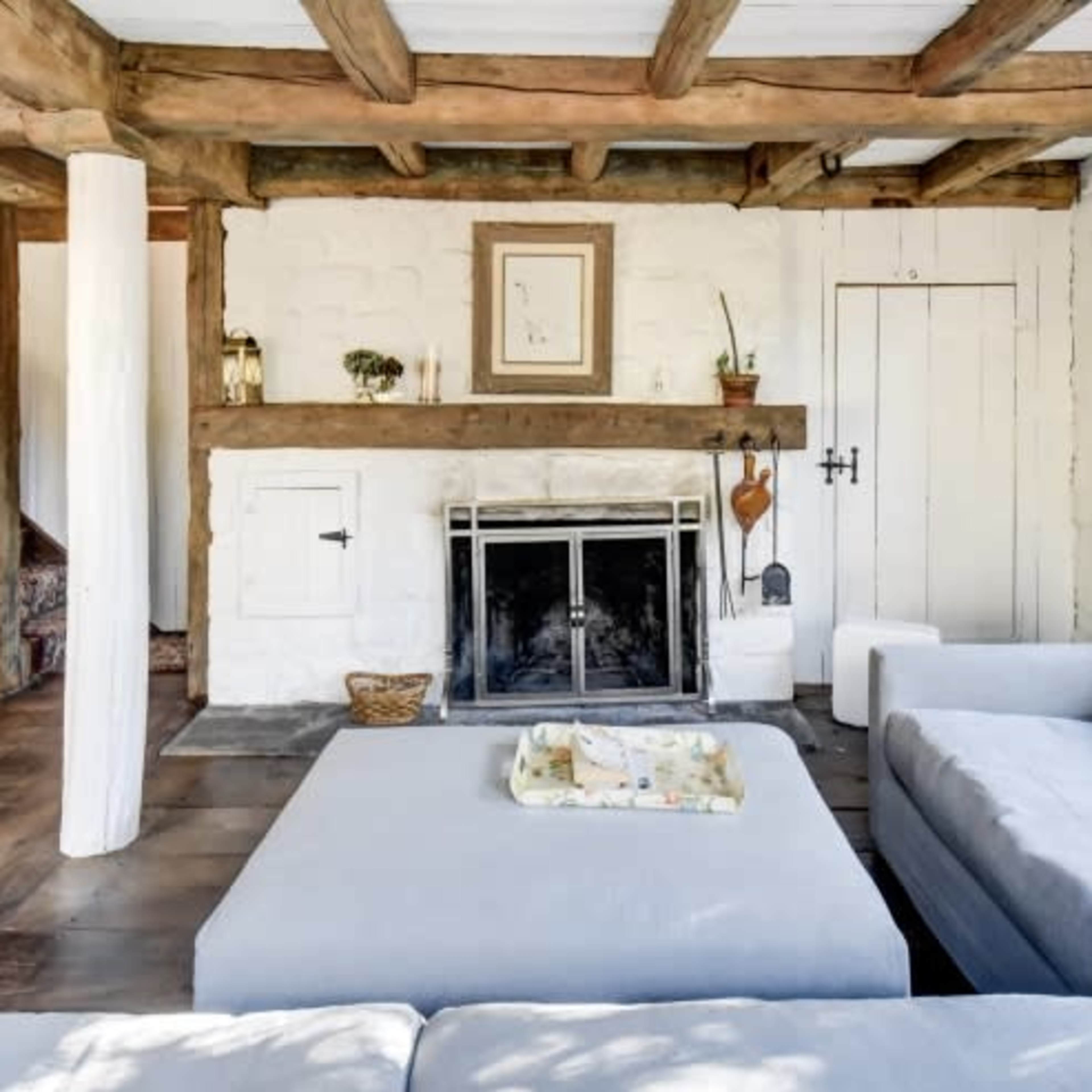 A cozy living room with exposed wooden beams, a stone fireplace, and a light-colored sofa arranged around a square coffee table.