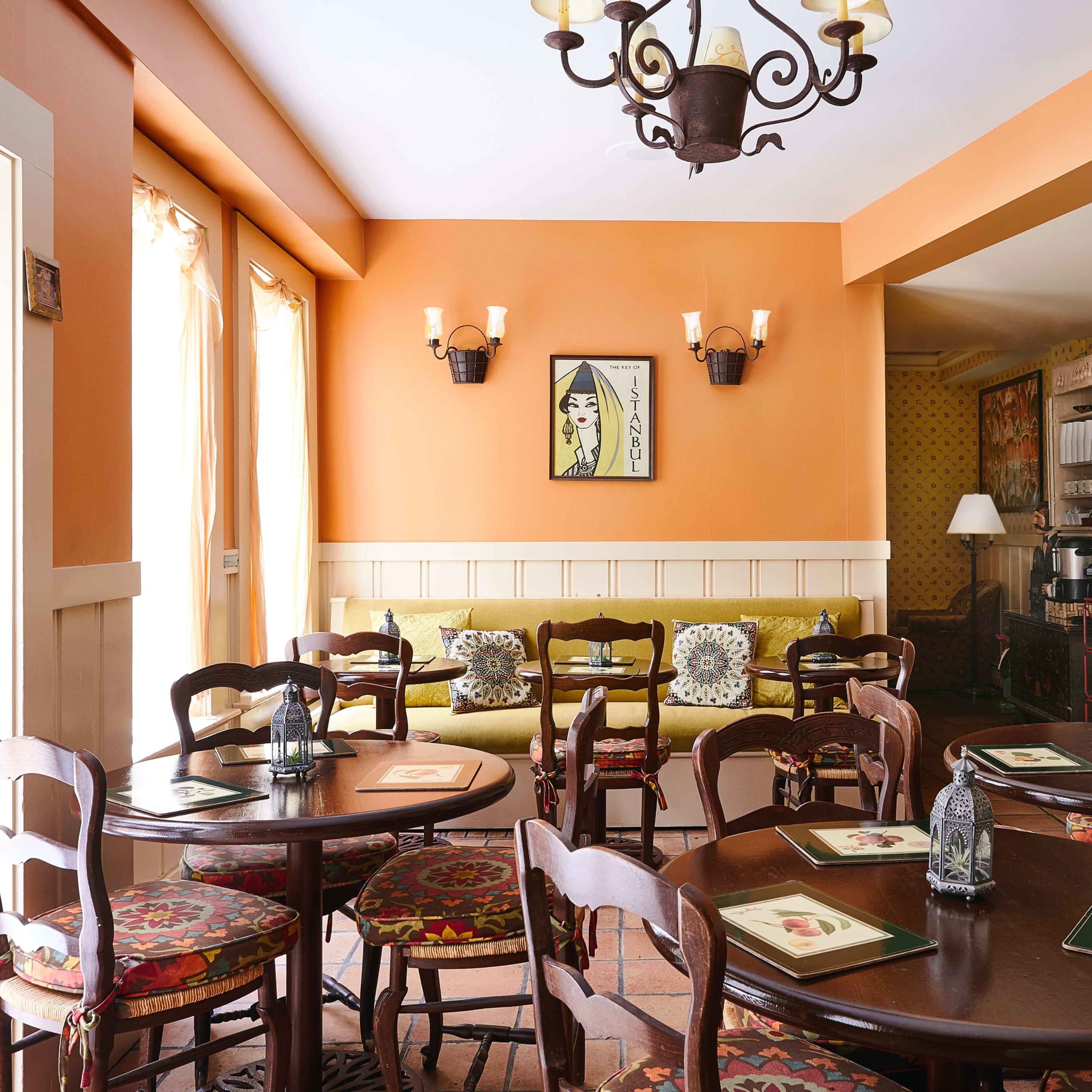 A cozy dining area with brown wooden tables and chairs, an orange wall, and a small seating nook against the wall, illuminated by natural light.