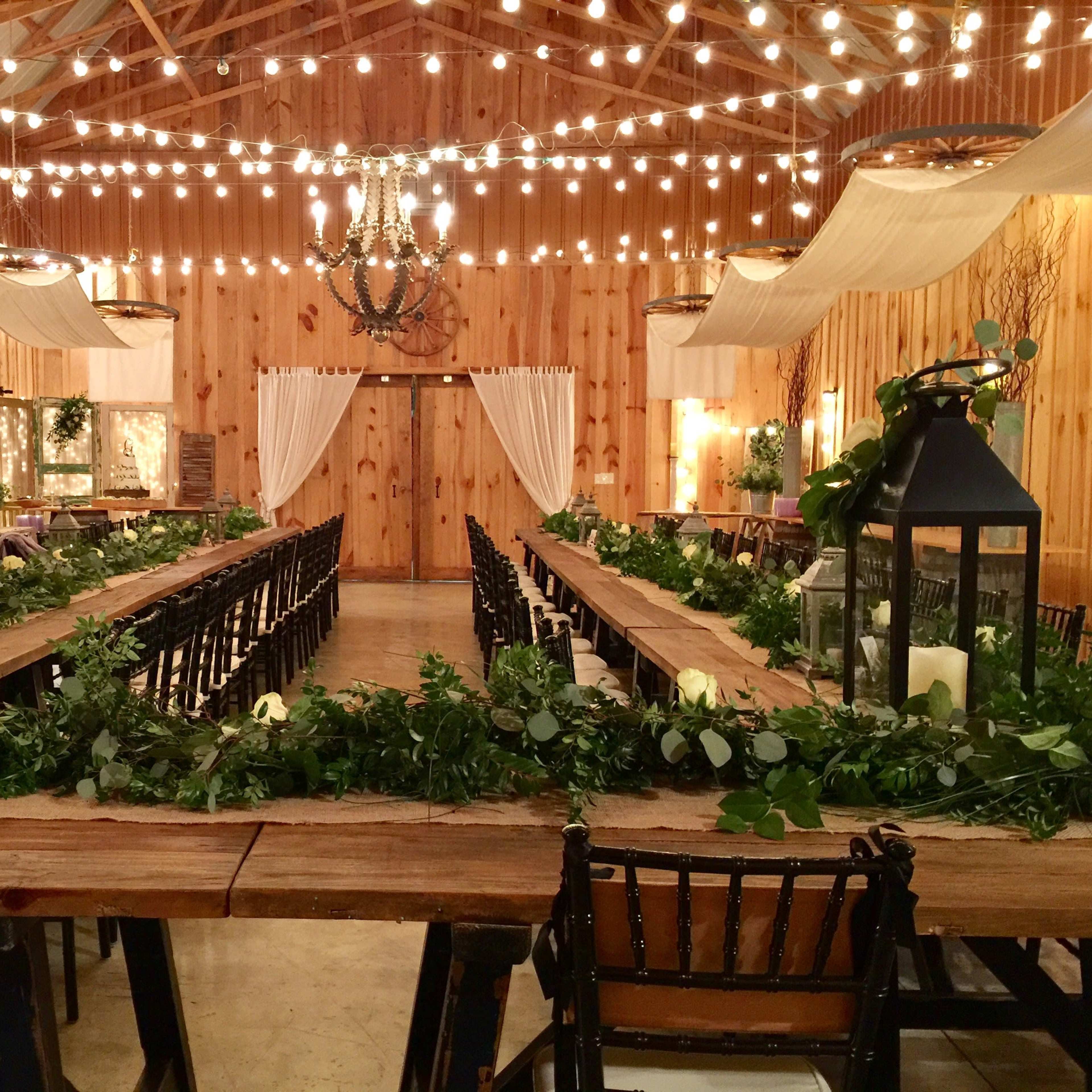 A spacious, wooden banquet hall is decorated with string lights and greenery along long wooden tables, with rows of dark chairs facing the center.