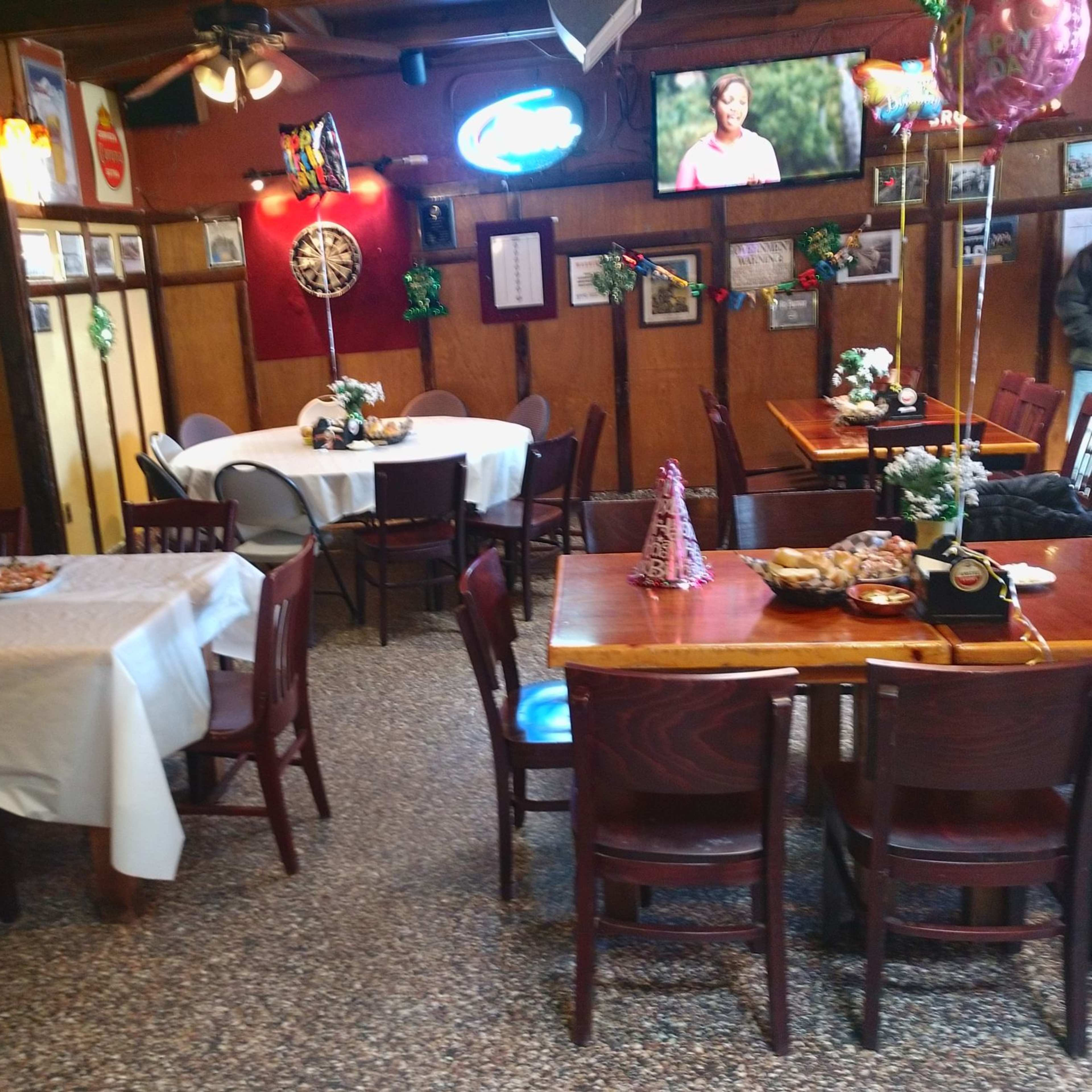 A cozy restaurant interior with wooden tables, chairs, and festive decorations, including balloons and a television screen.