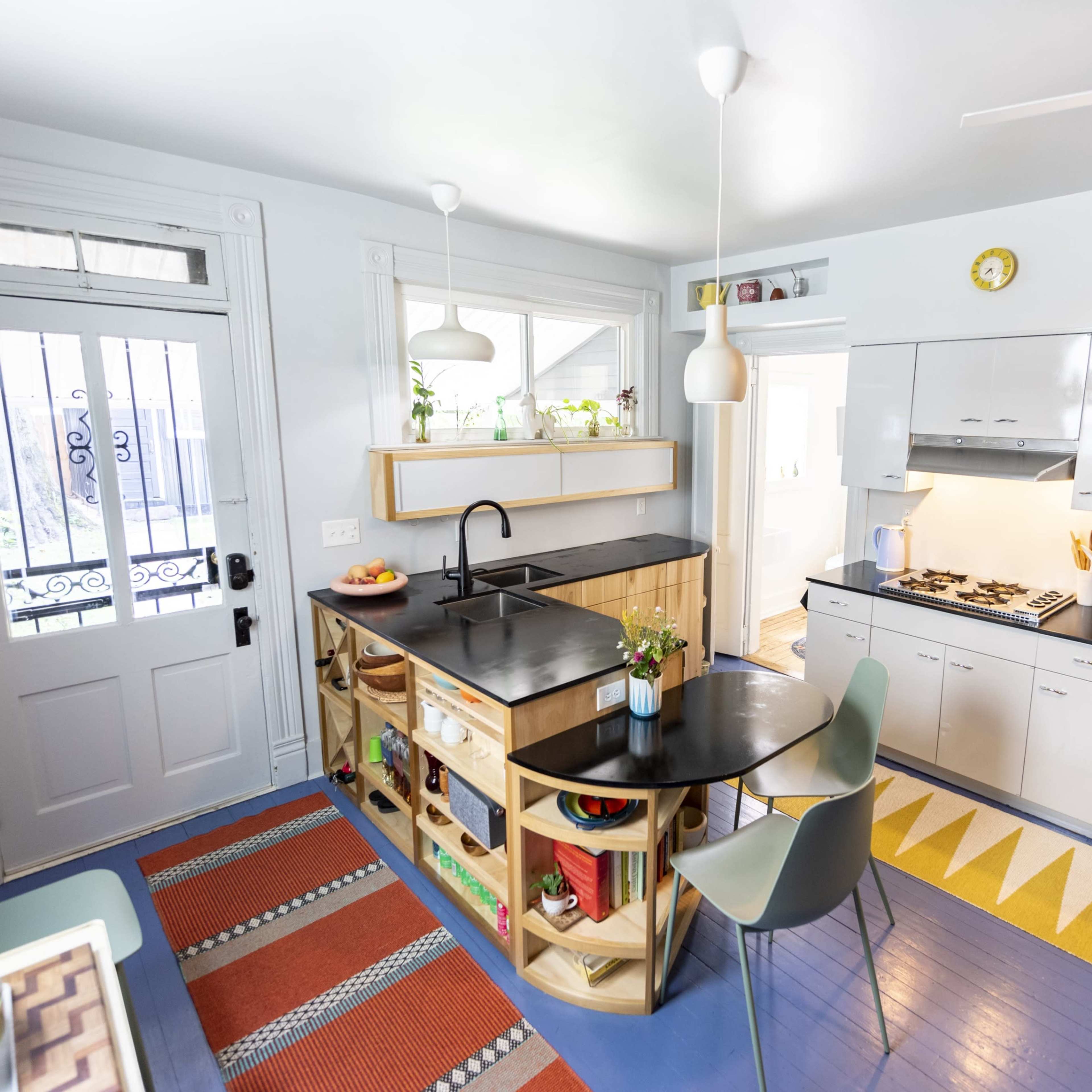 A modern kitchen with a black countertop, open shelving, and colorful flooring, featuring a round table and a door with a decorative grill.