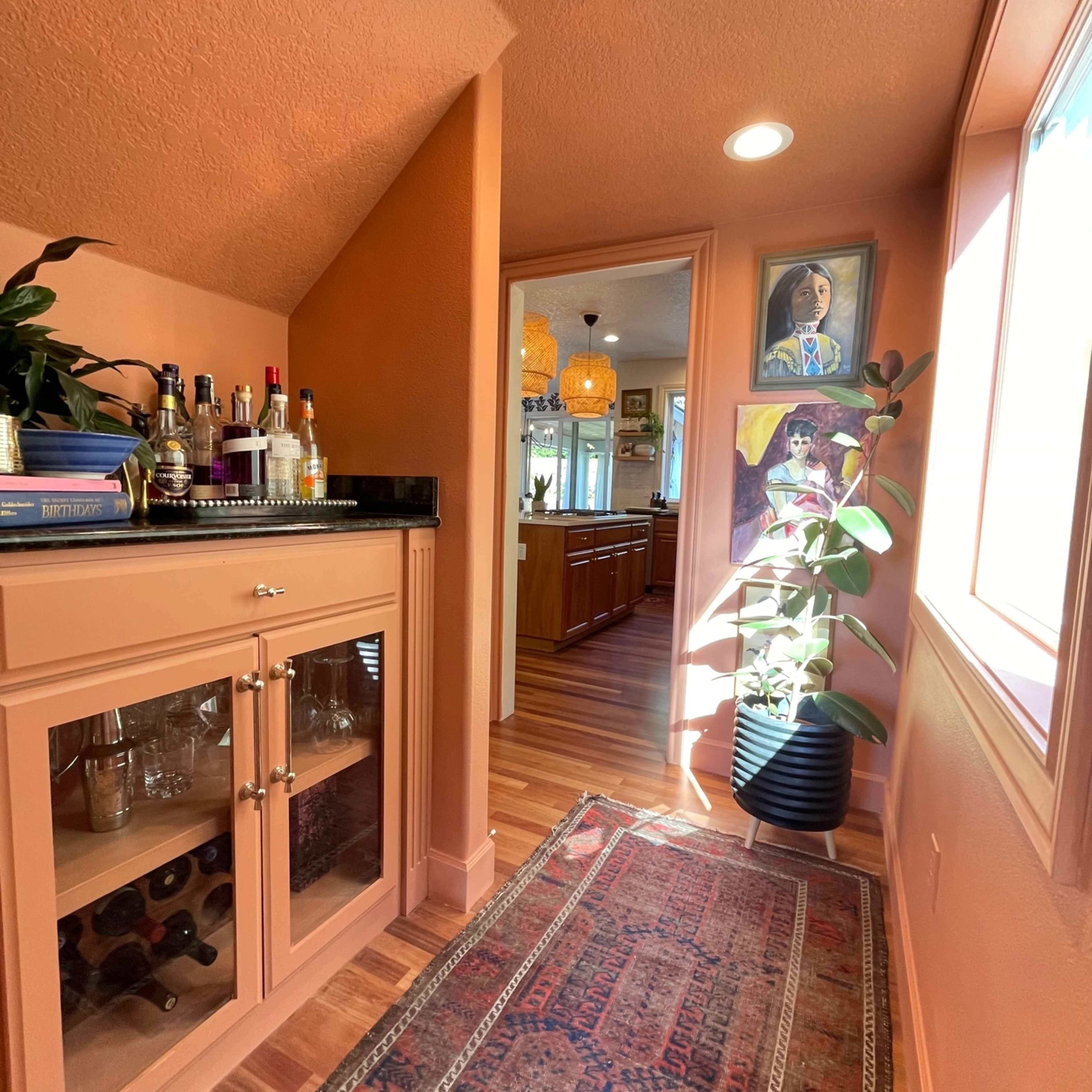 A narrow hallway features a bar cabinet with glassware, a potted plant, and artwork on the walls, leading to a kitchen area beyond.