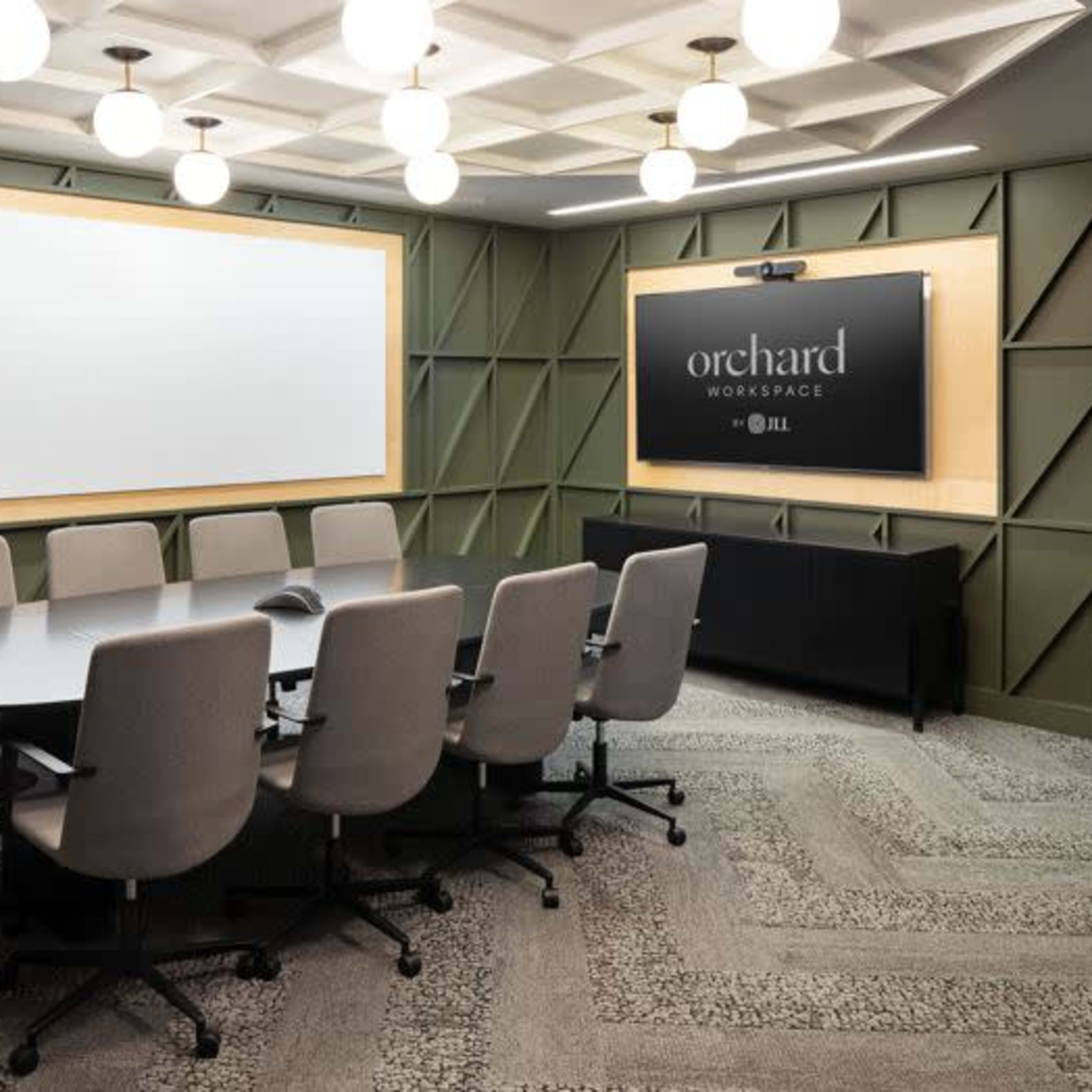 A conference room features a circular table surrounded by ergonomic chairs, two blank whiteboards, and a display screen on a textured green wall.
