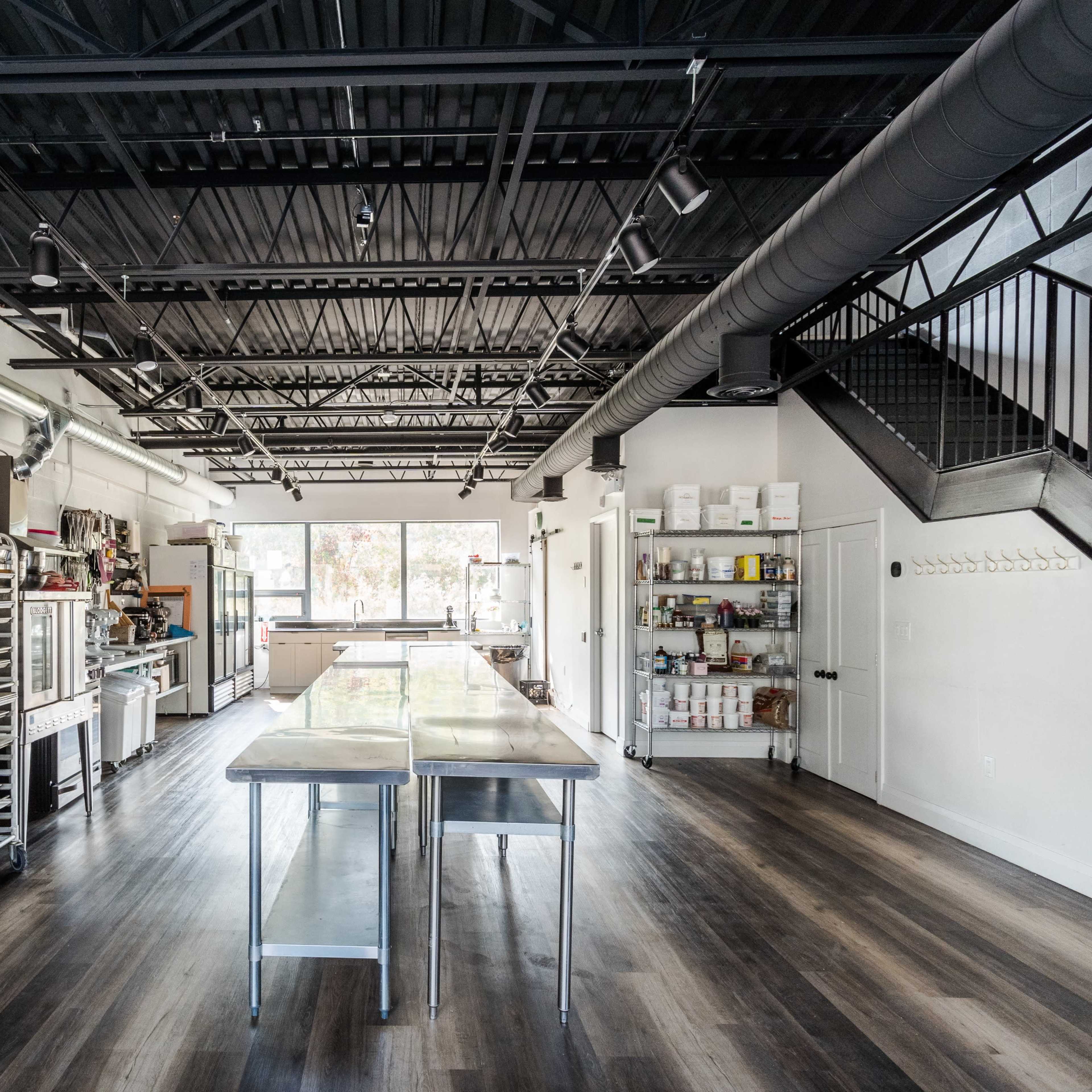 A modern commercial kitchen with stainless steel tables, various kitchen equipment, and a staircase leading to an upper level.