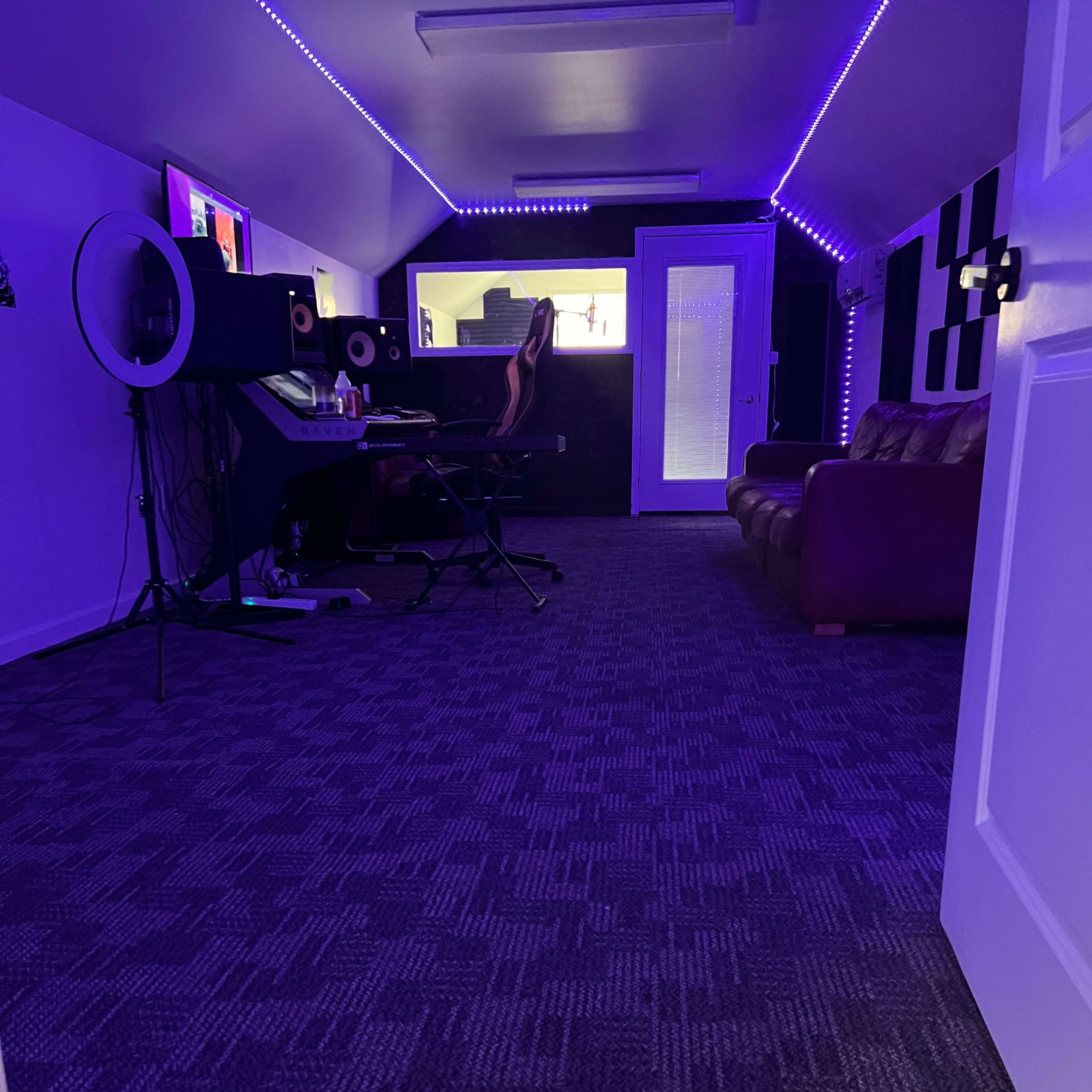 A room with purple lighting features a computer setup, a microphone, and a brown couch, all placed on a carpeted floor.