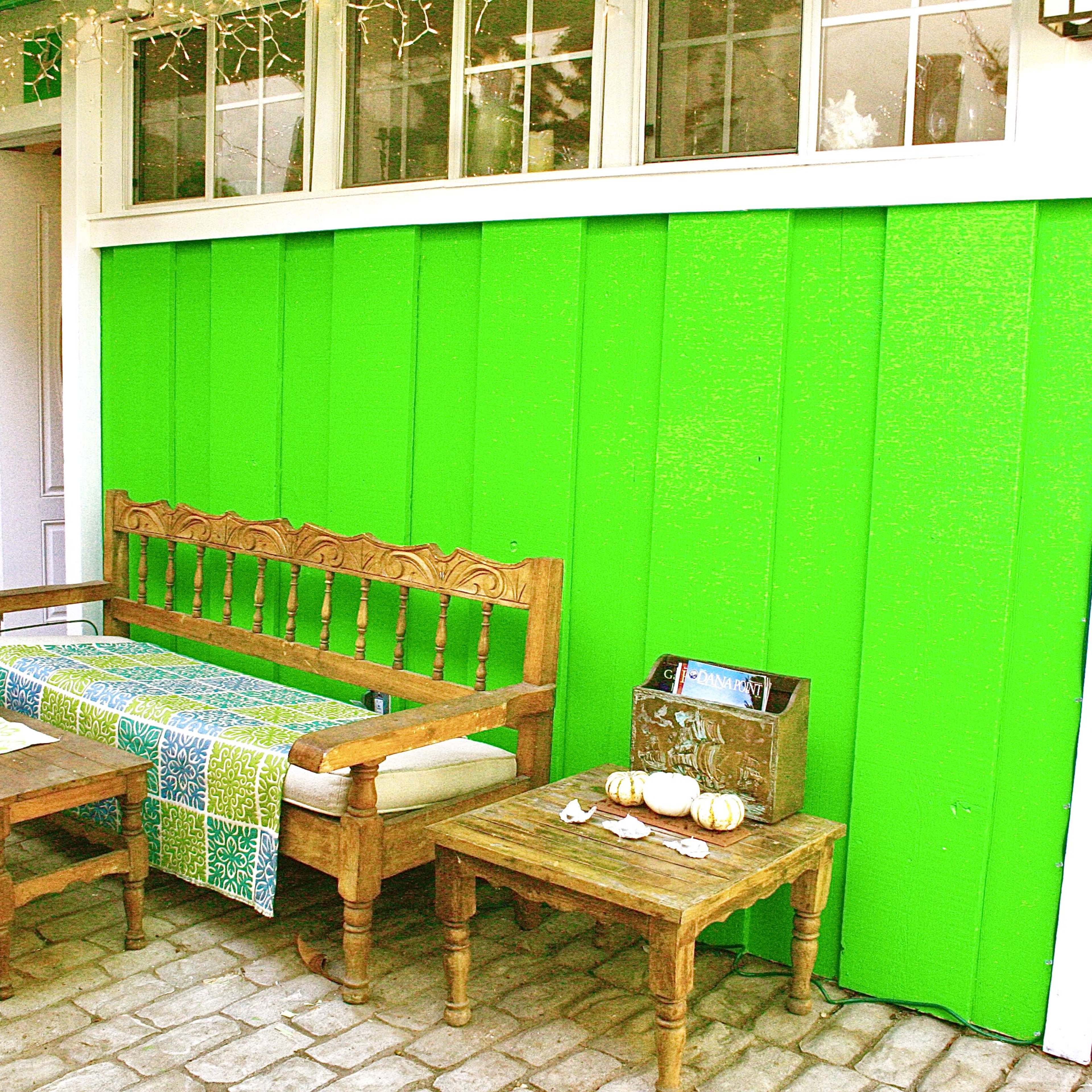 A wooden bench and two tables sit in front of a vibrant green wall, with a patterned cloth draped over the bench and small decorative items on the tables.