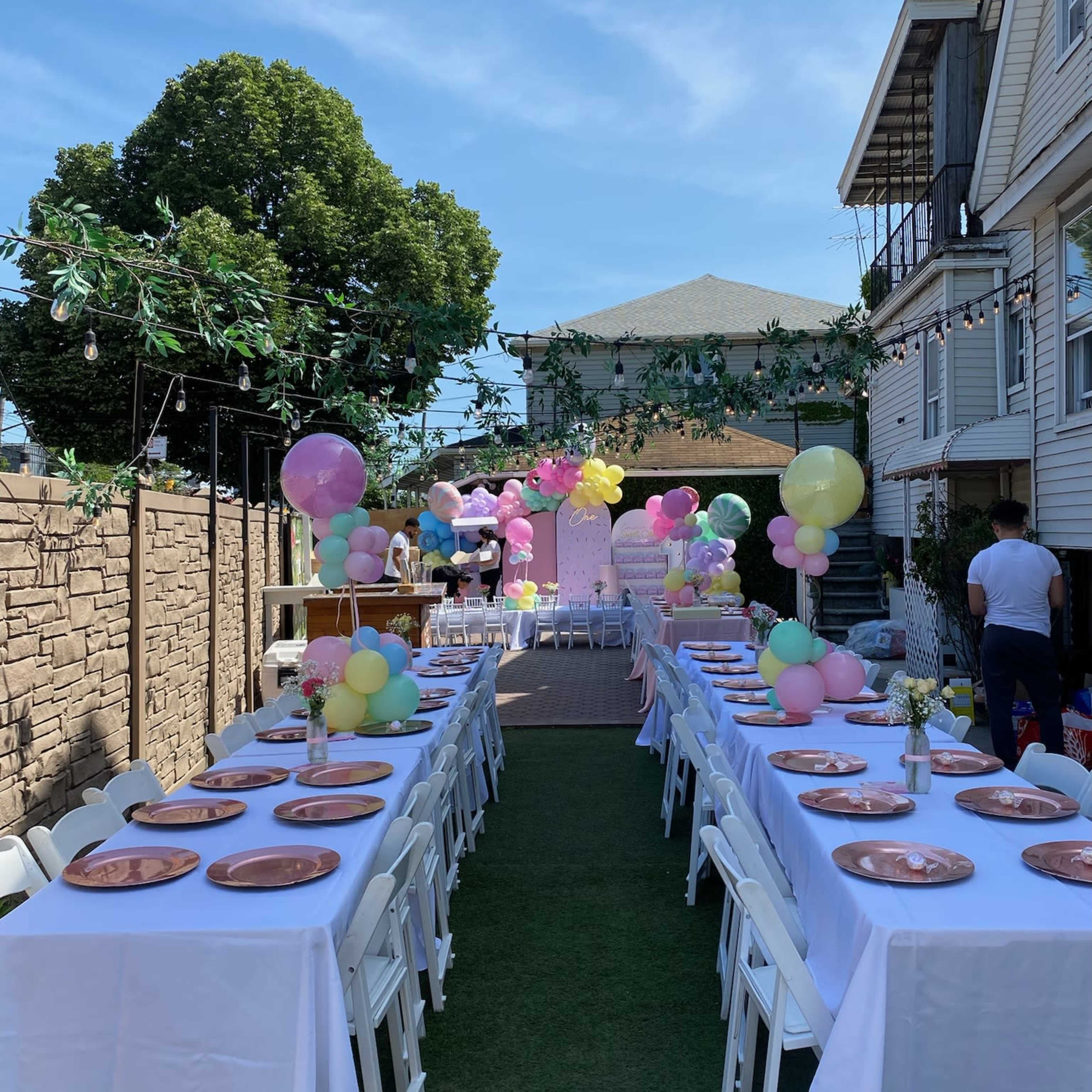 A decorated outdoor space features long tables with white tablecloths set up for a celebration, surrounded by colorful balloons and greenery.