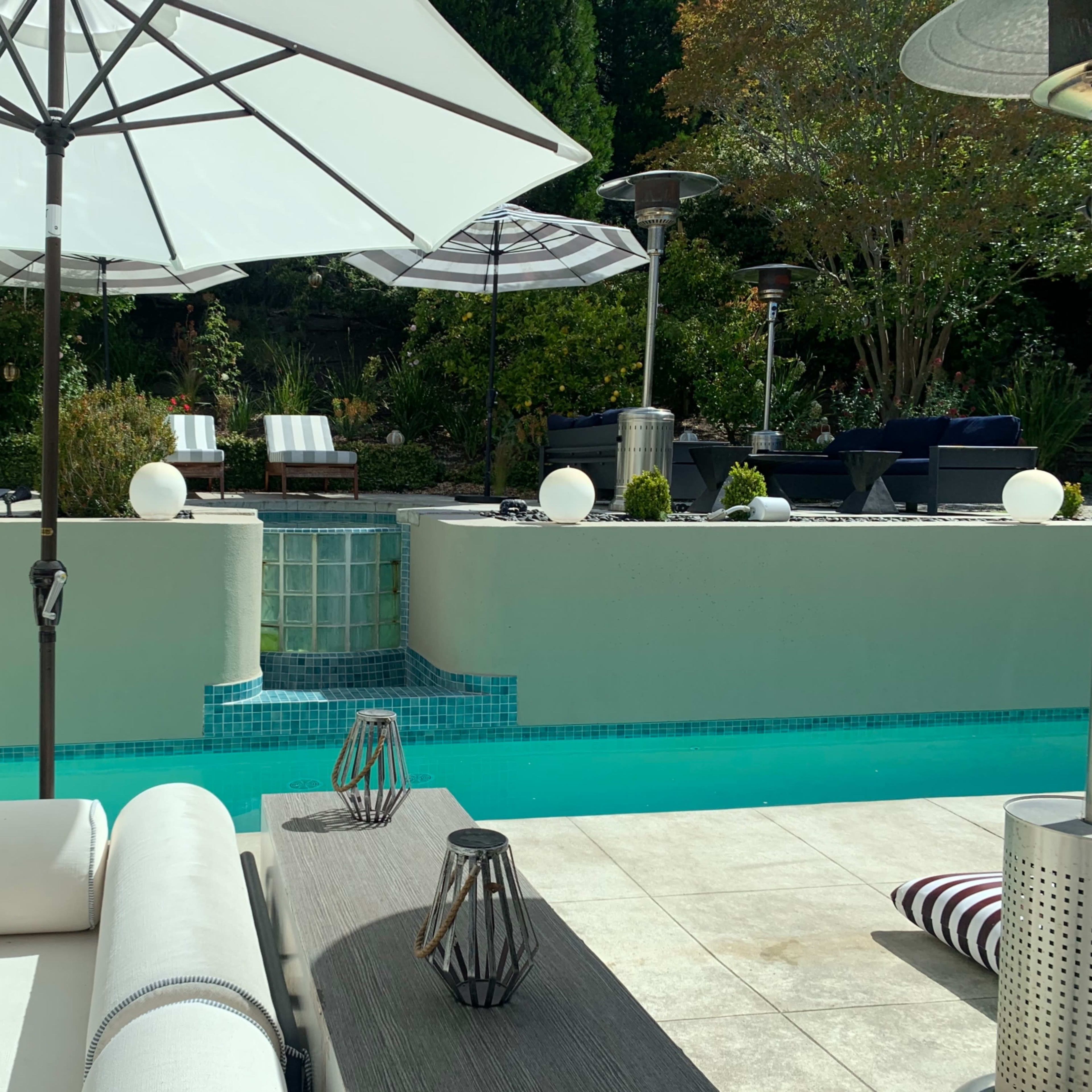 A modern outdoor pool area with lounge chairs, tables, and patio umbrellas surrounded by greenery.
