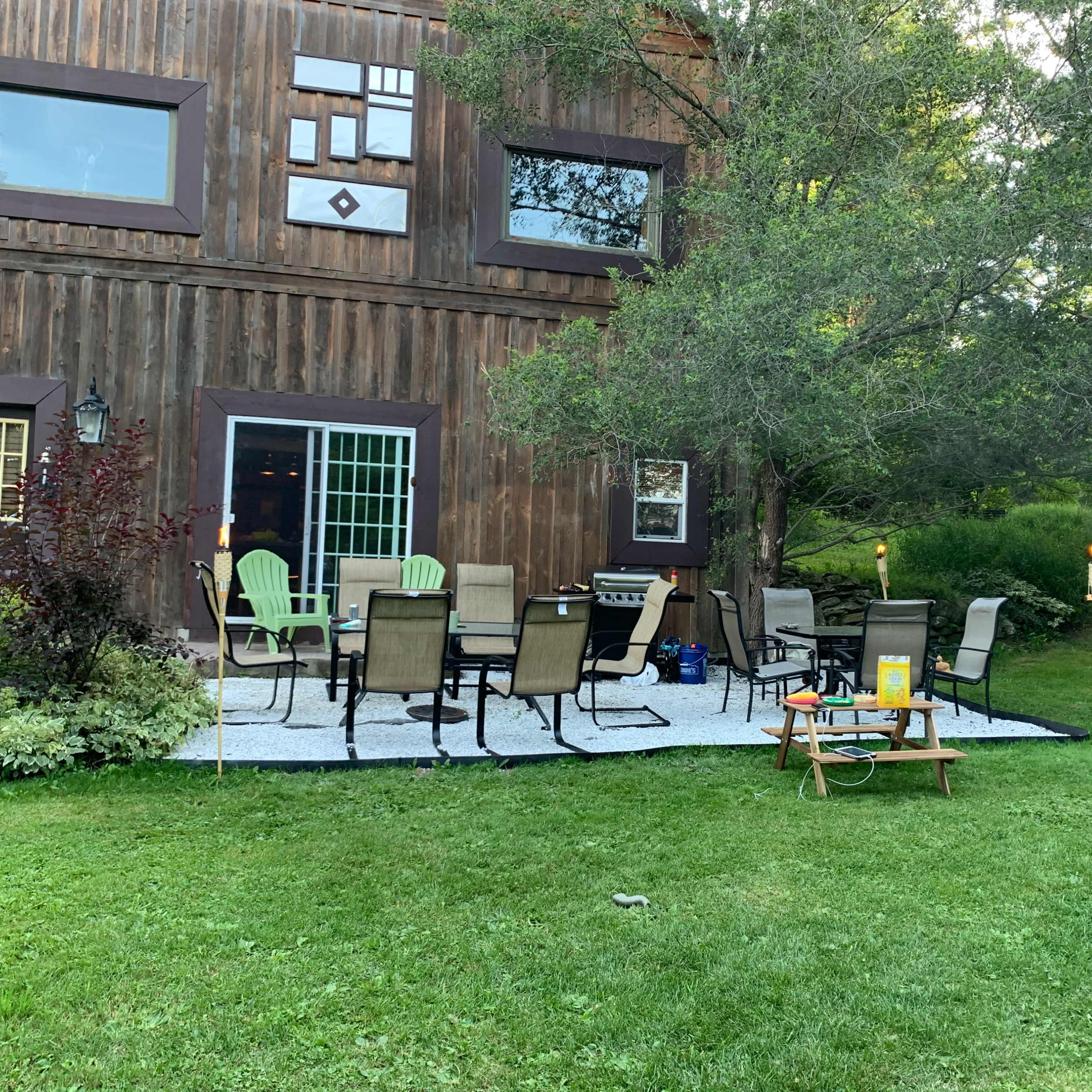 A wooden building with a green door has a gravel seating area surrounded by chairs and a table, situated in a grassy yard.