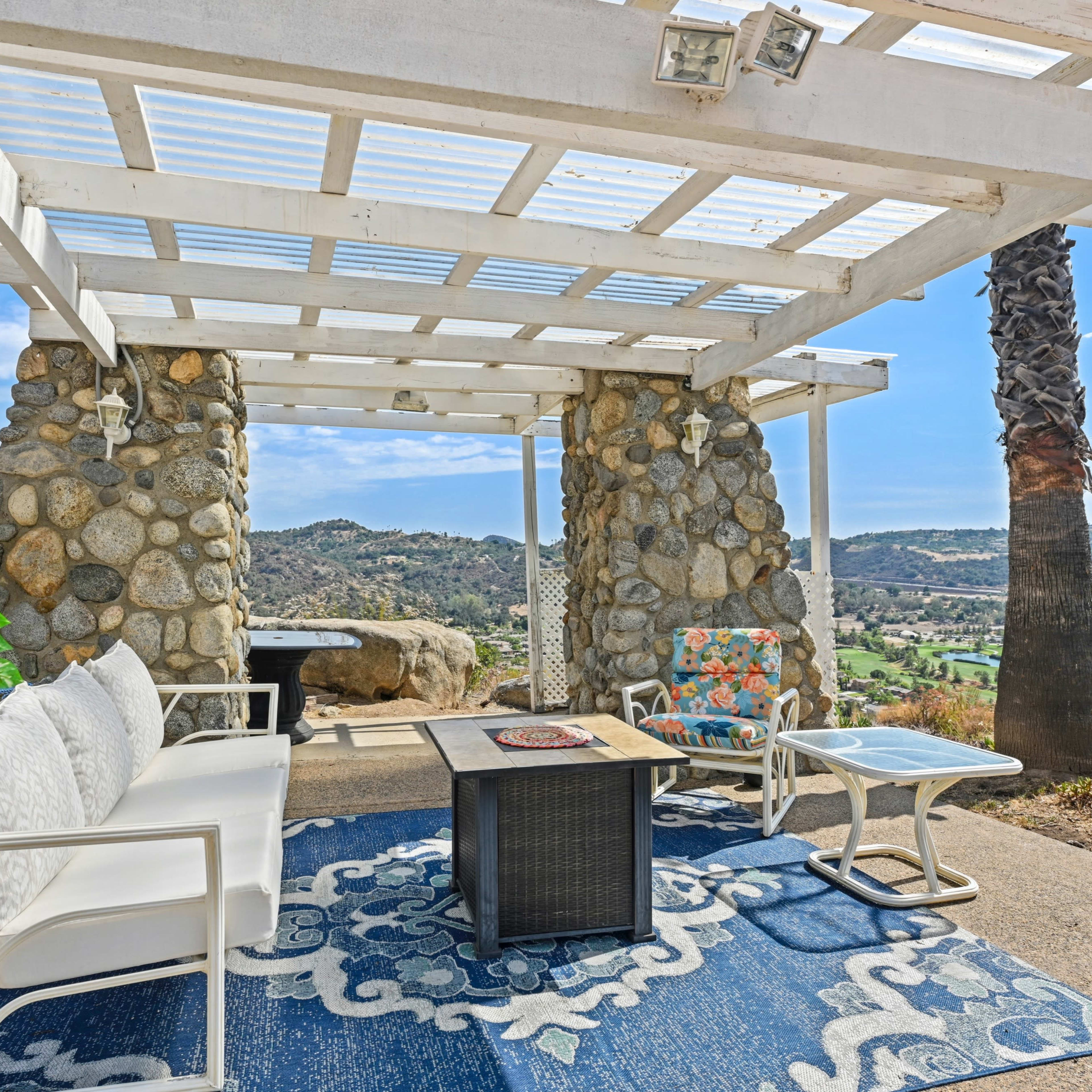 A patio area with a stone structure, white seating, and a table, set against a scenic backdrop of hills and valleys.