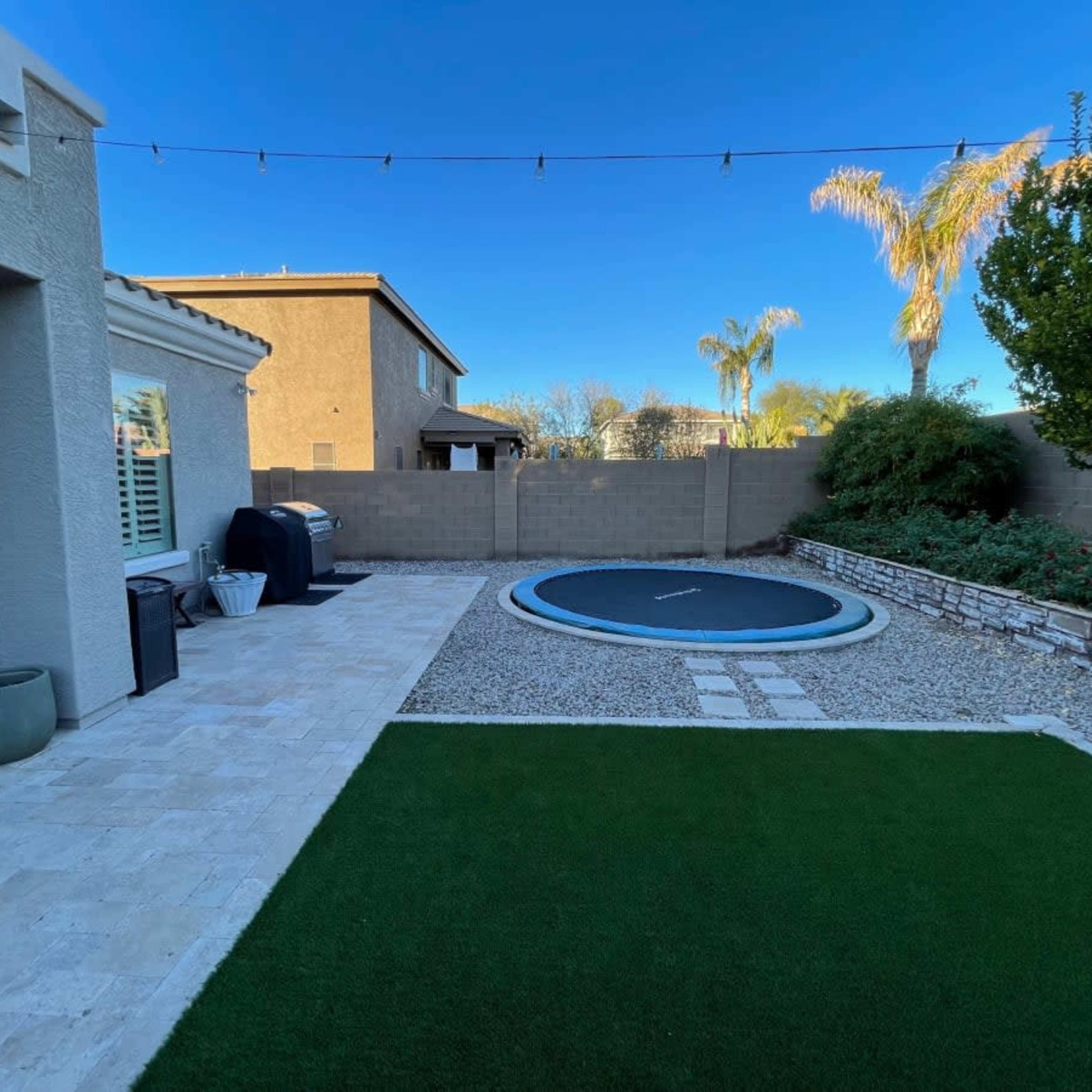 A backyard with a trampoline, artificial grass, a stone path, and a barbecue grill, surrounded by a low wall and several palm trees.