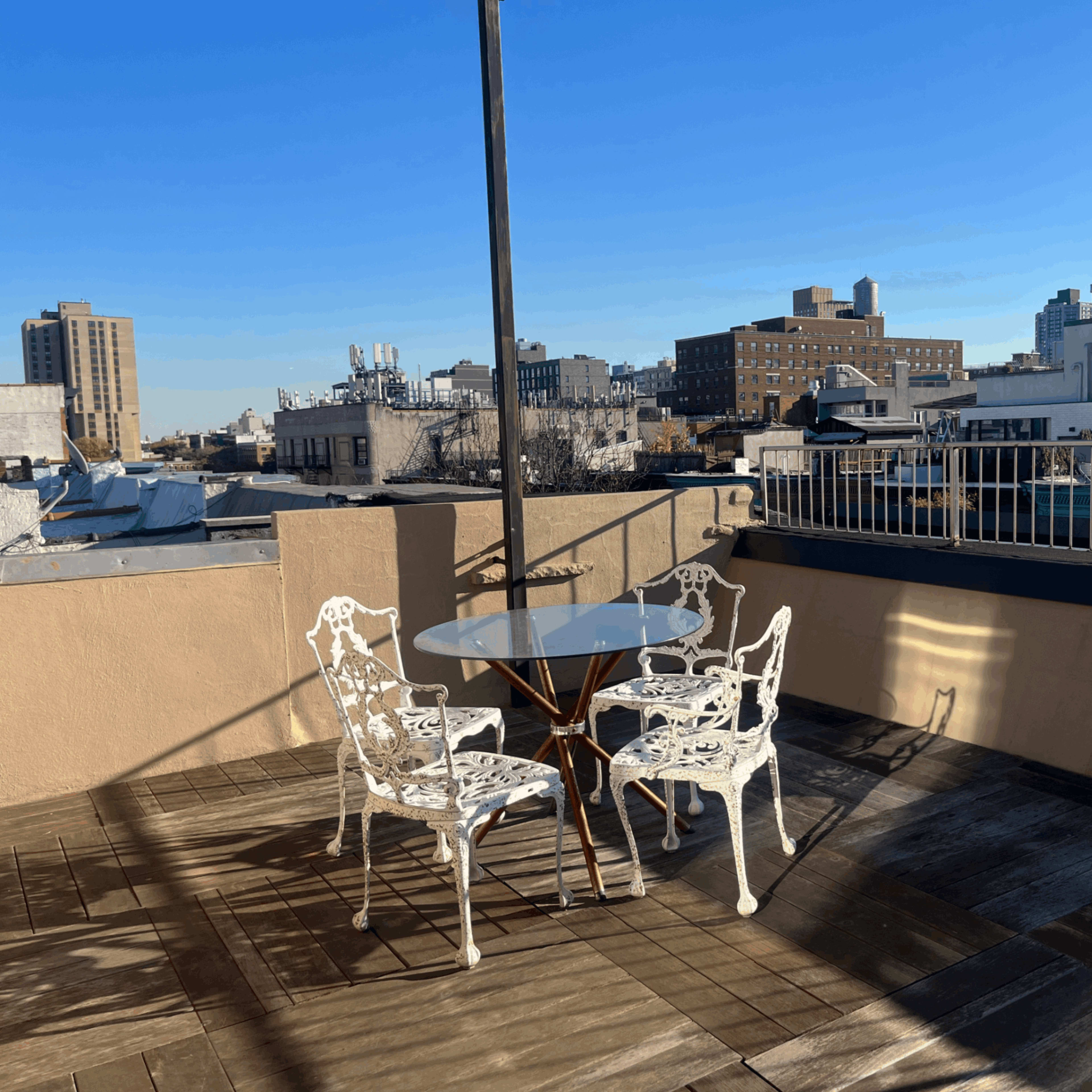 A rooftop patio with a glass table and four white metal chairs set against a backdrop of urban buildings and a clear blue sky.