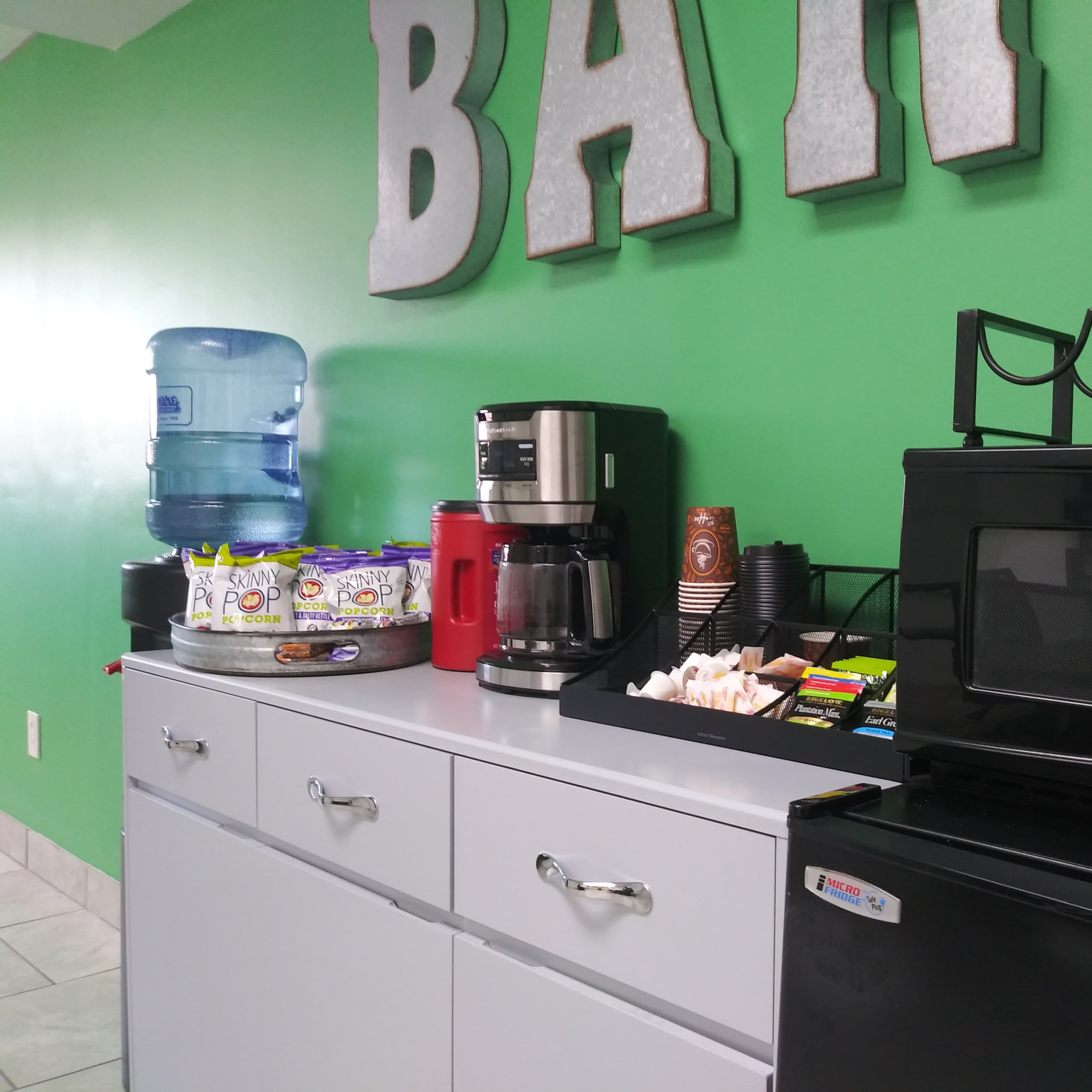 A kitchenette area features a water cooler, coffee maker, microwave, and assorted snacks on a countertop in front of a green wall with a sign that says "BAR."