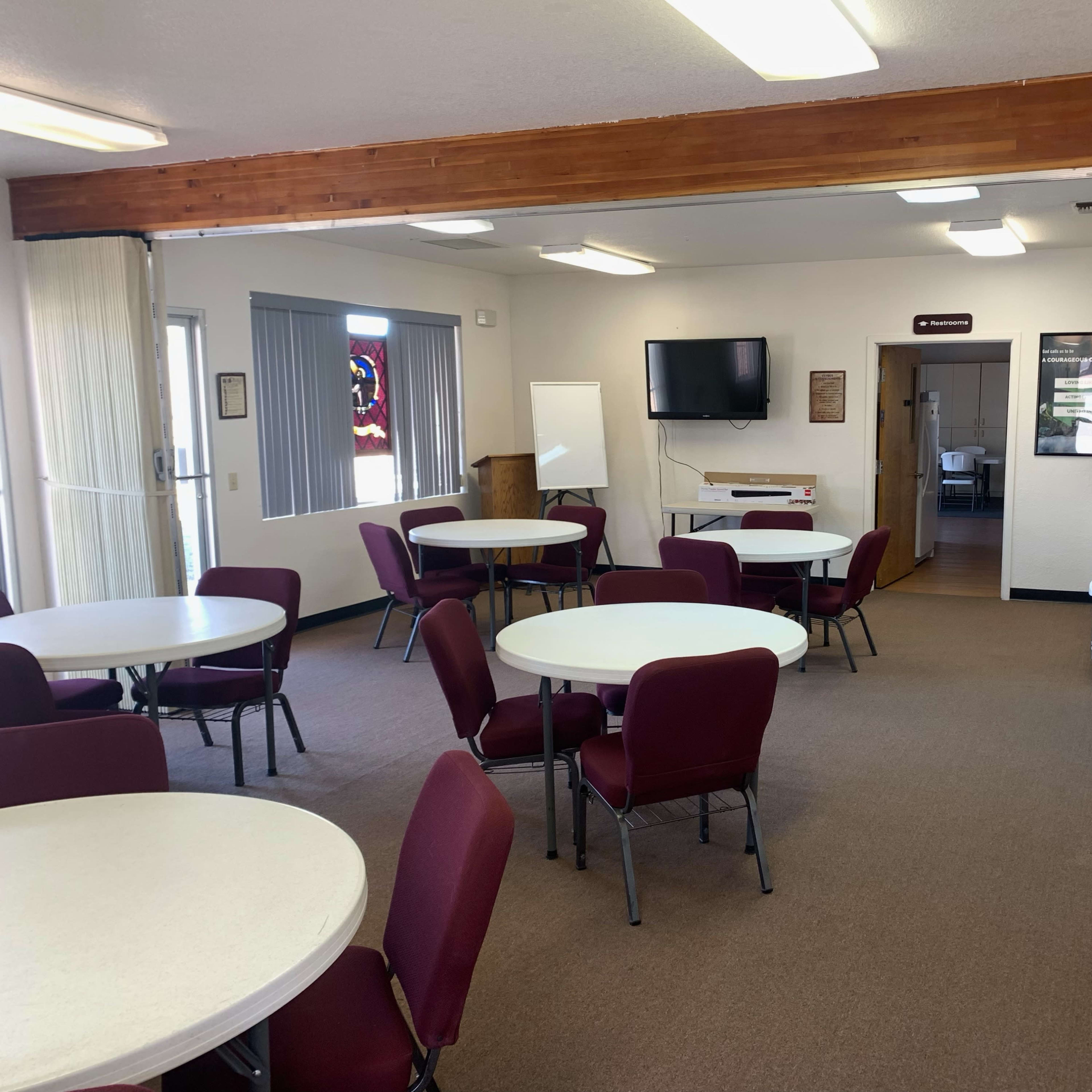 A room with several round tables and maroon chairs, a television mounted on the wall, and a kitchen area in the background.