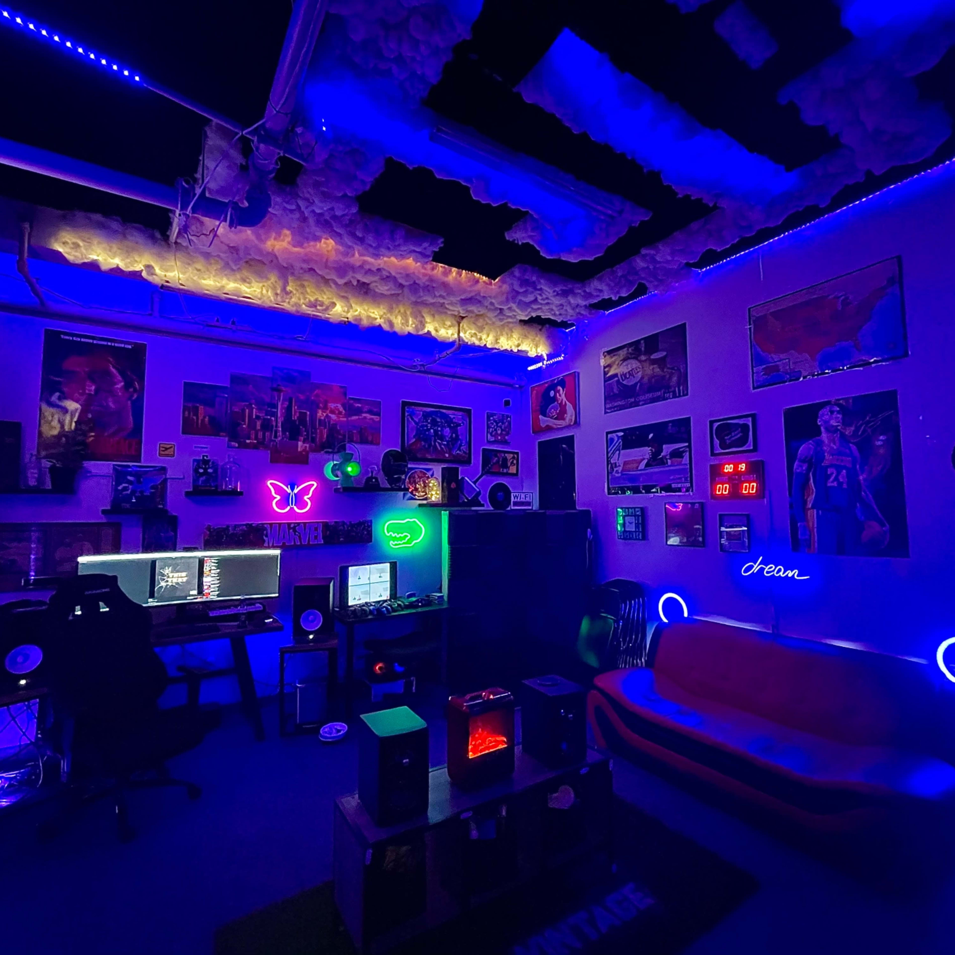 A dimly lit room decorated with colorful neon lights, a computer setup with multiple monitors, vinyl records on the walls, and a couch.