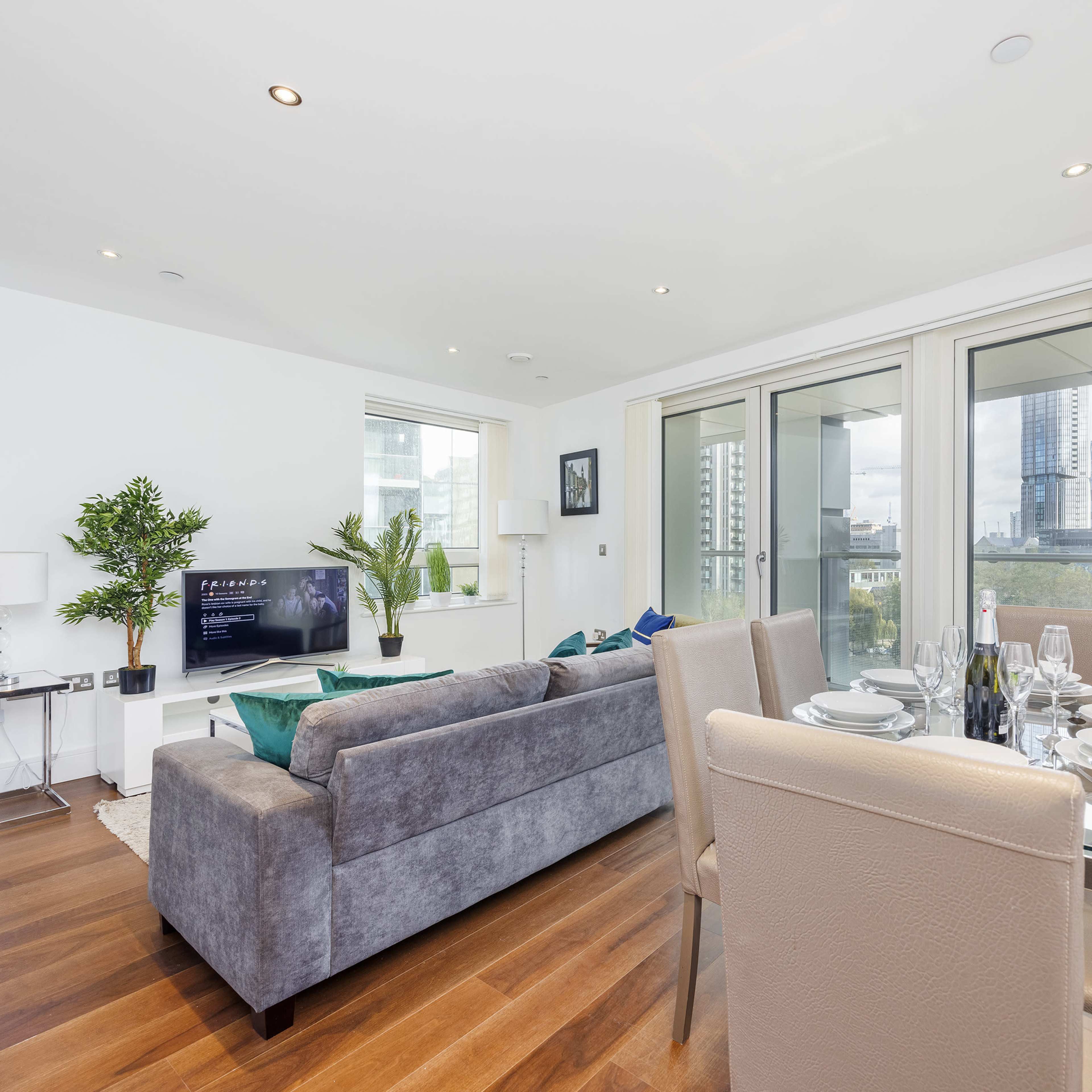 A modern living area with a gray sofa, a dining table set for four, and large windows providing a view of a city skyline.