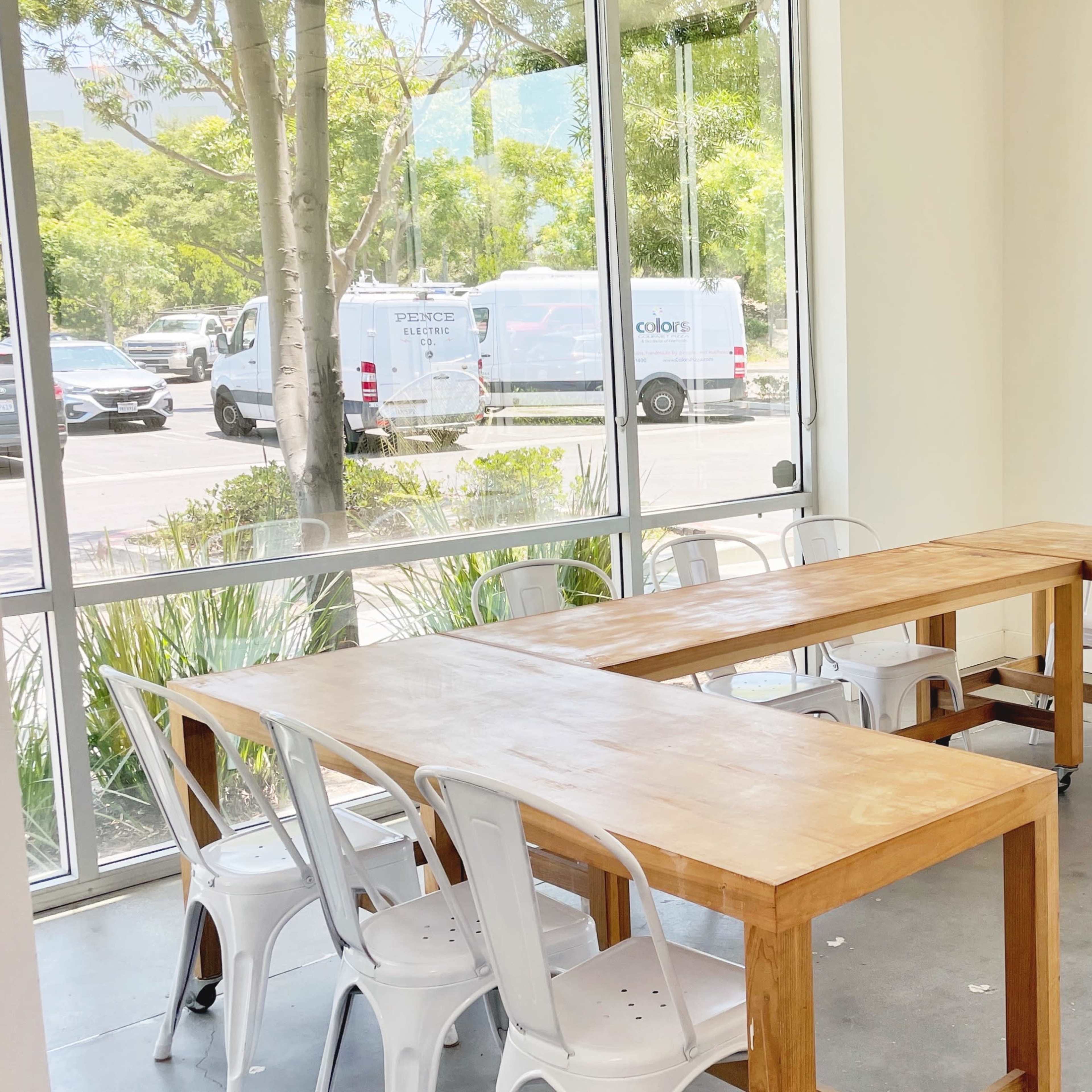 A bright room features two wooden tables arranged in an L-shape, surrounded by several white chairs, with large windows revealing greenery and parked vehicles outside.