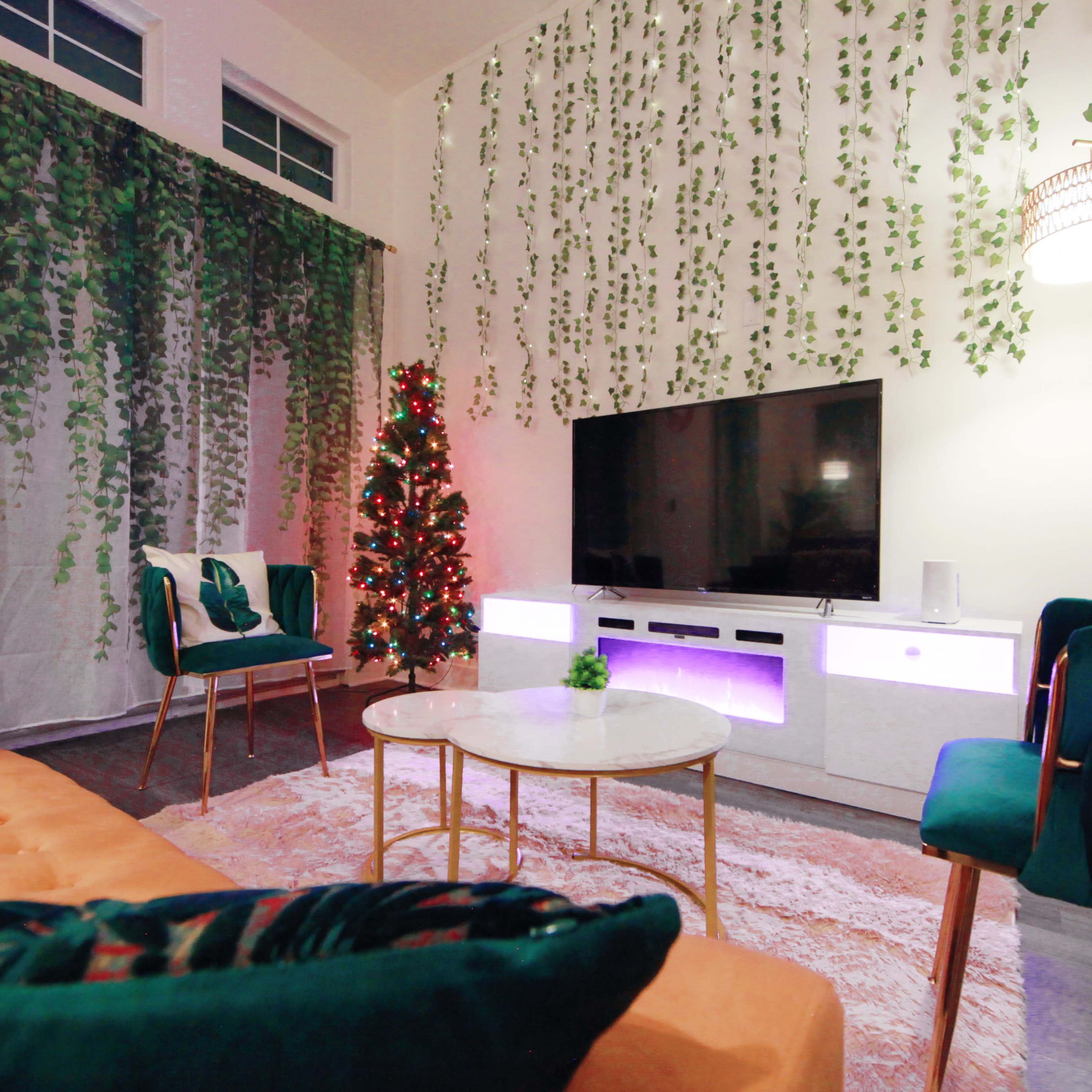 The living room features a television mounted on a white cabinet, surrounded by green plants and a small decorated Christmas tree, with modern furniture arranged around a low coffee table.