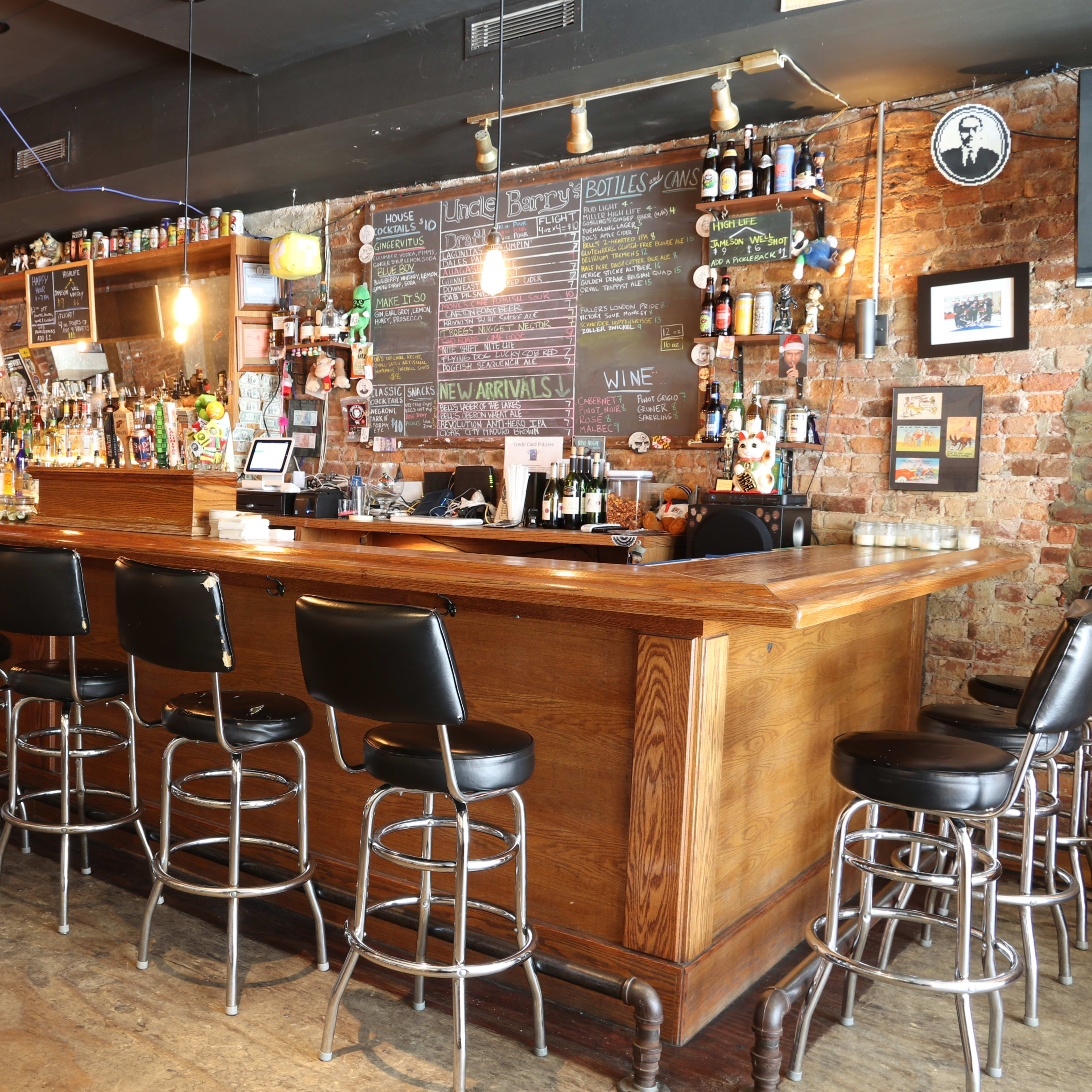 A spacious bar with a wooden counter lined with black stools, surrounded by exposed brick walls and shelves filled with various bottles of alcohol.