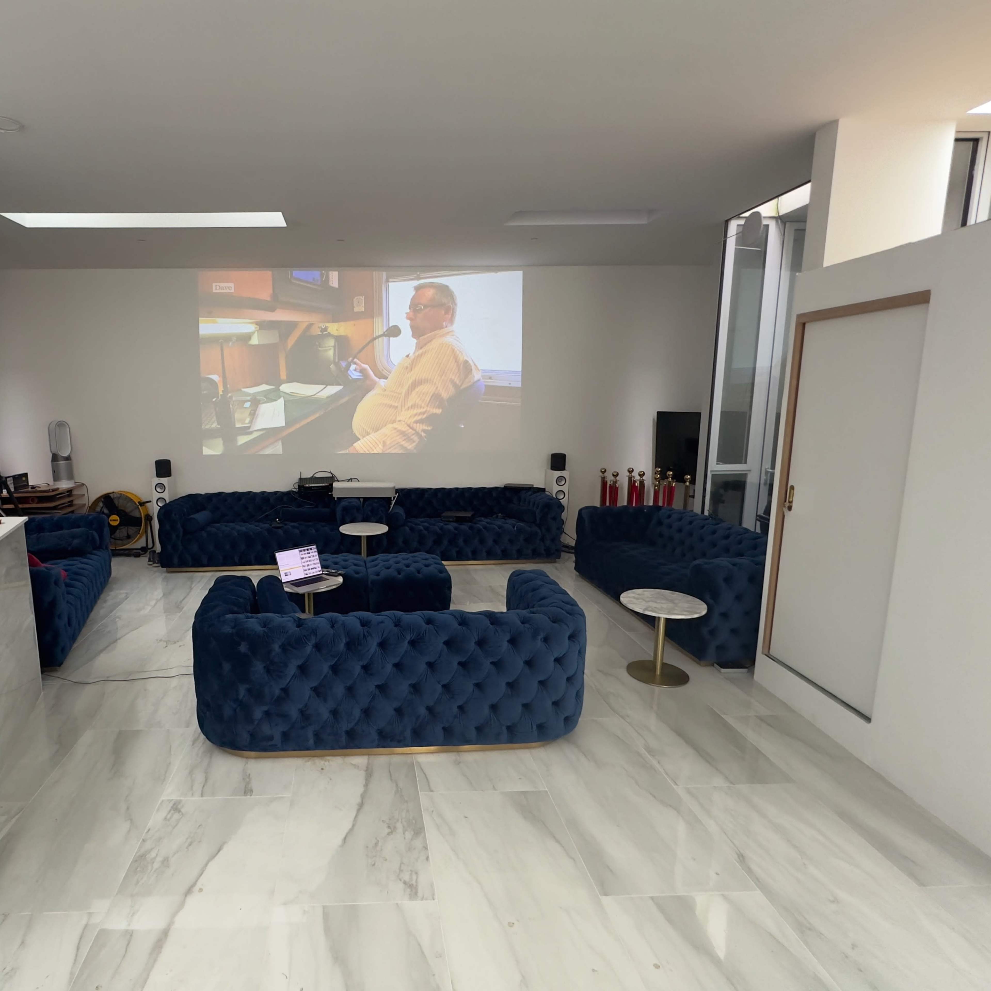 A modern lounge area features blue tufted sofas, a small round table, and a projector screen displaying a seated person.