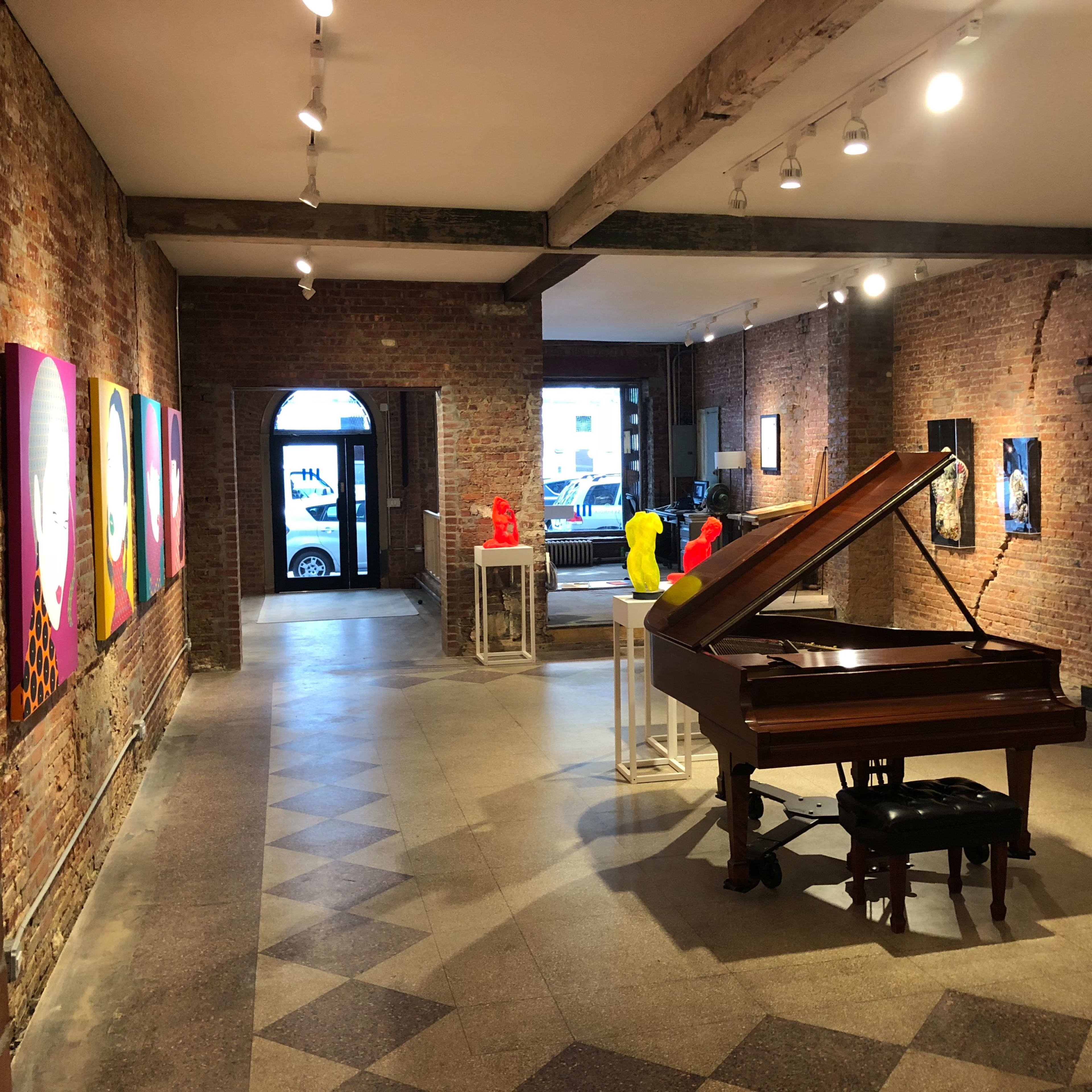 A spacious art gallery with exposed brick walls featuring colorful artworks, a grand piano in the foreground, and modern sculptures displayed on pedestals.