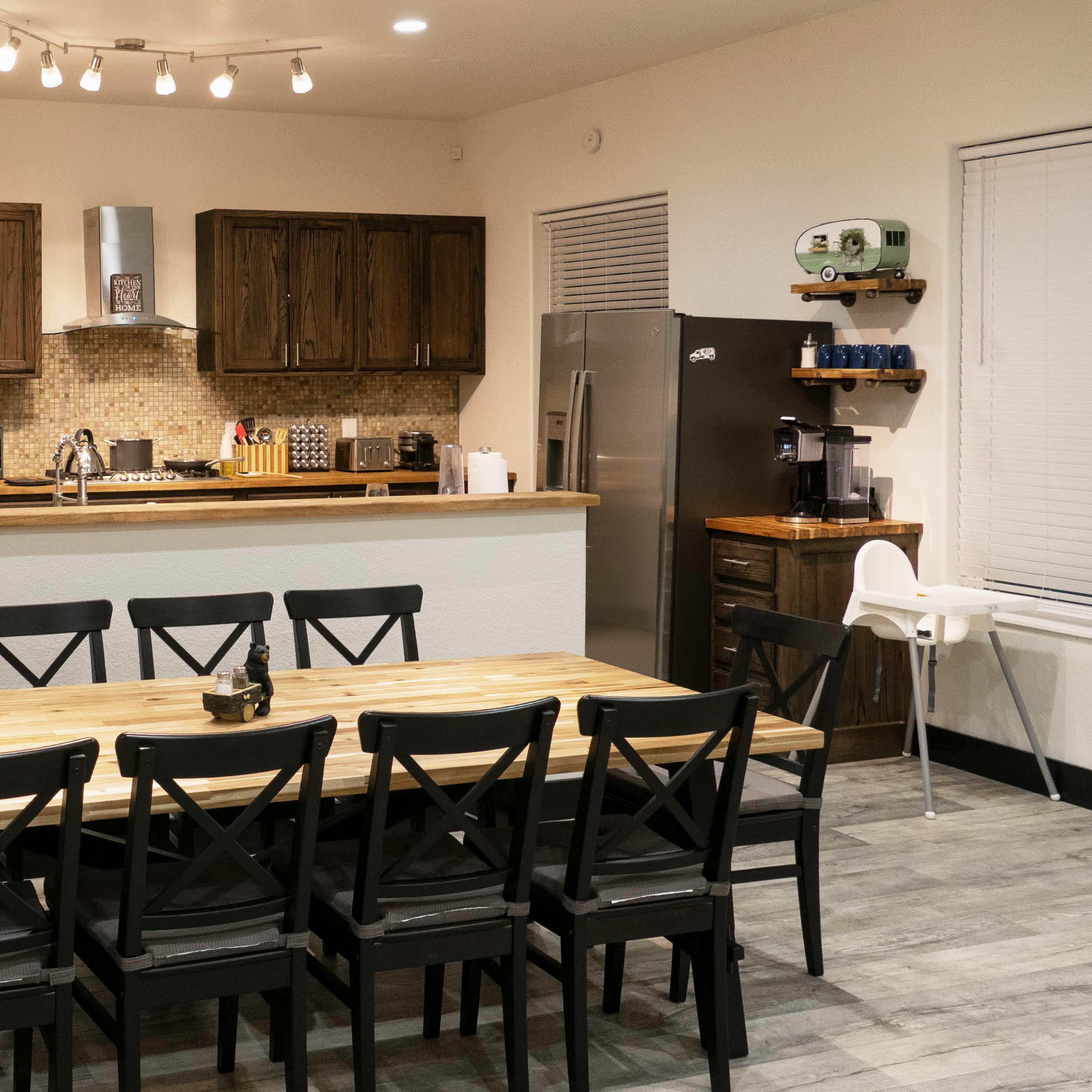 A spacious kitchen and dining area features a large wooden table surrounded by black chairs, with dark wooden cabinetry and modern appliances visible in the background.
