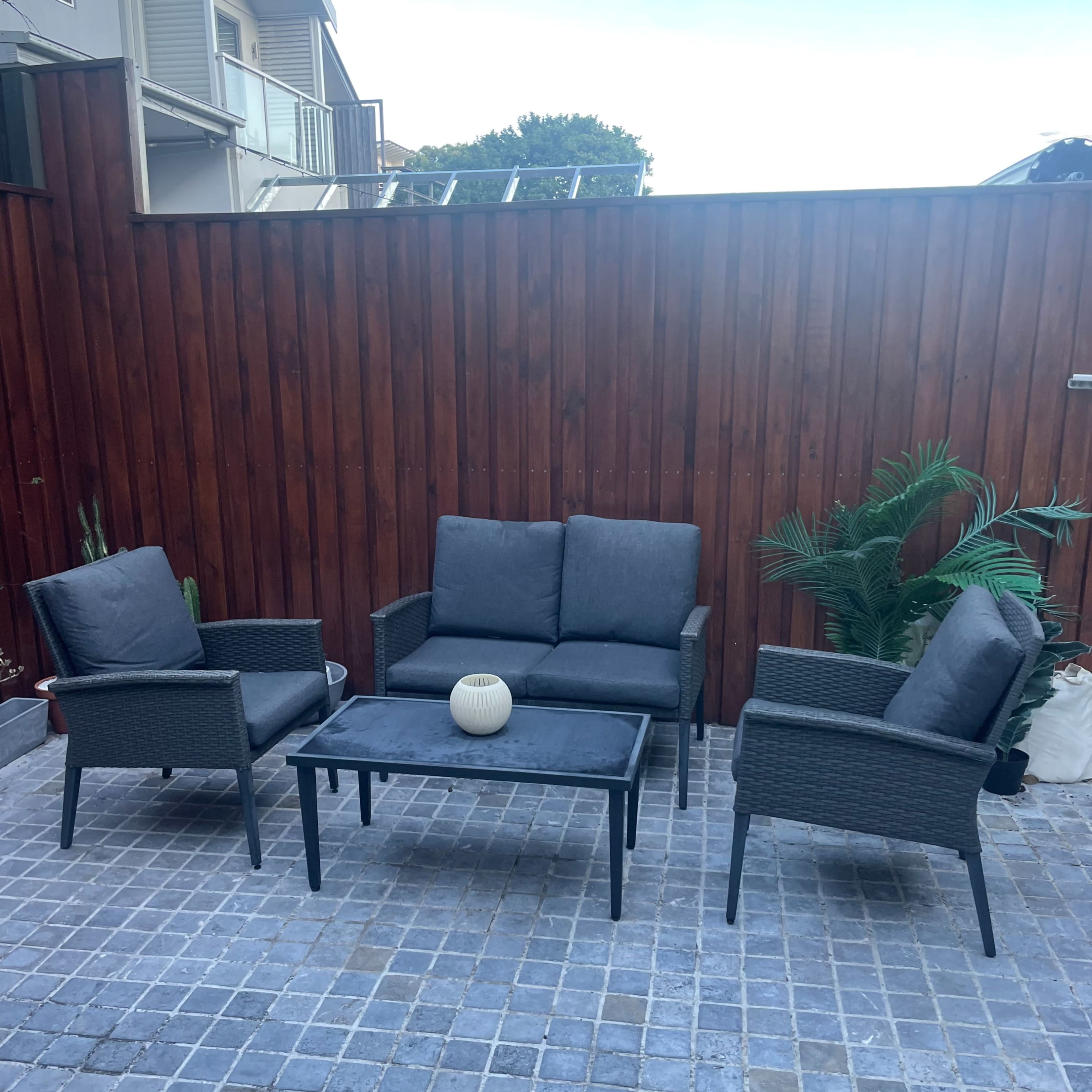 A patio area features a gray seating set with two chairs, a loveseat, and a table, surrounded by a wooden fence and potted plants.