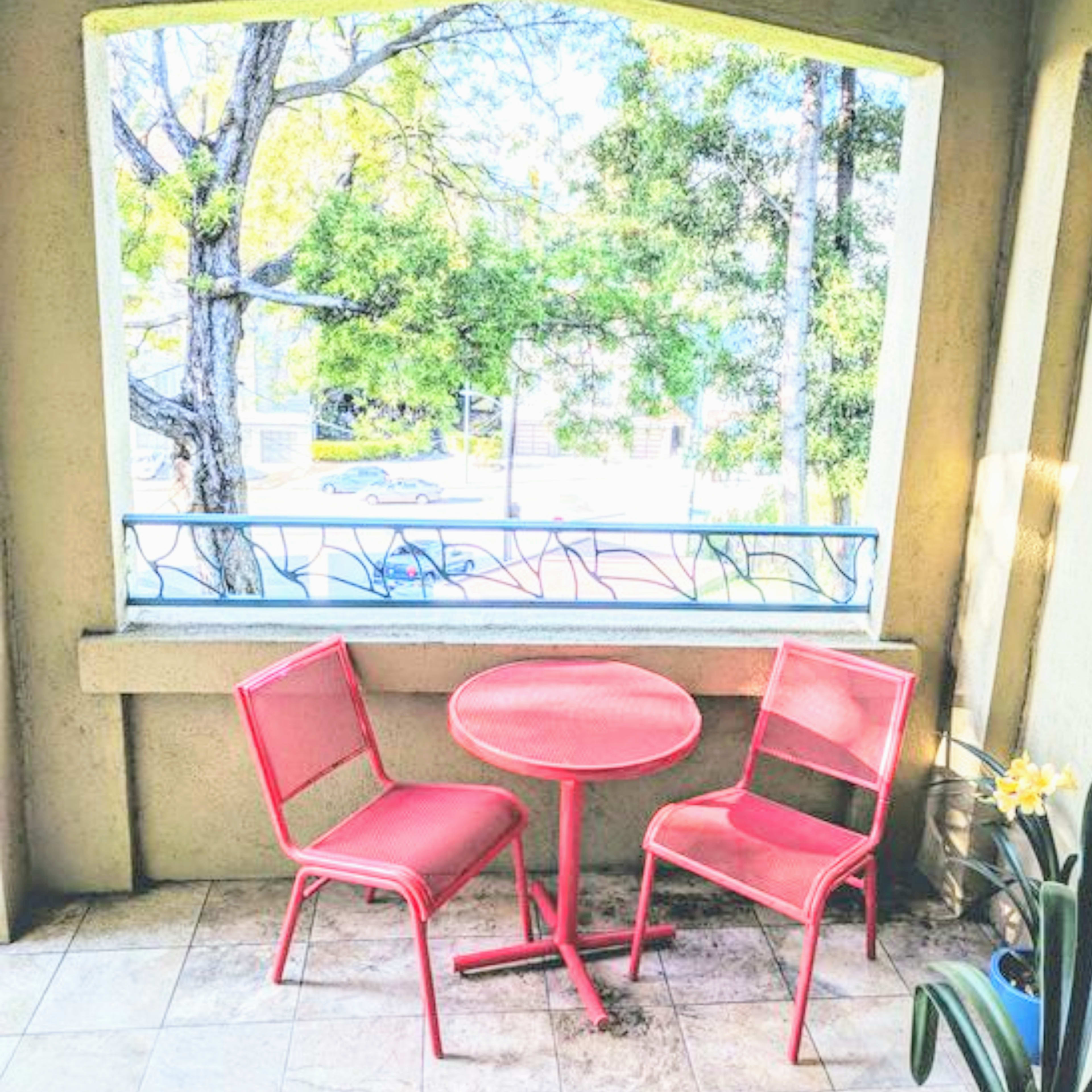 A small balcony features a round table and two red chairs beside a window framed by trees.