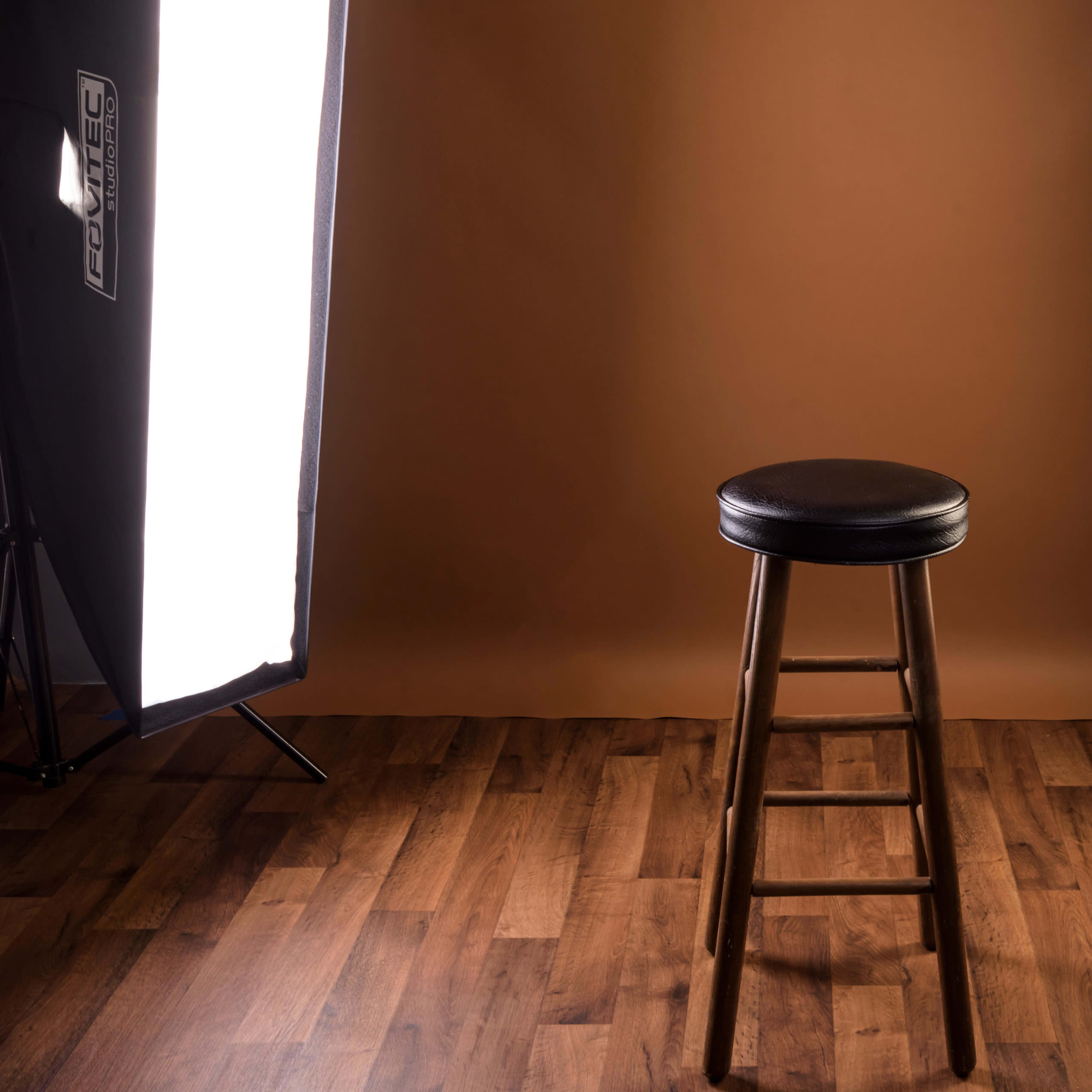 A black stool stands in the center of a well-lit studio with a brown backdrop and wooden flooring.