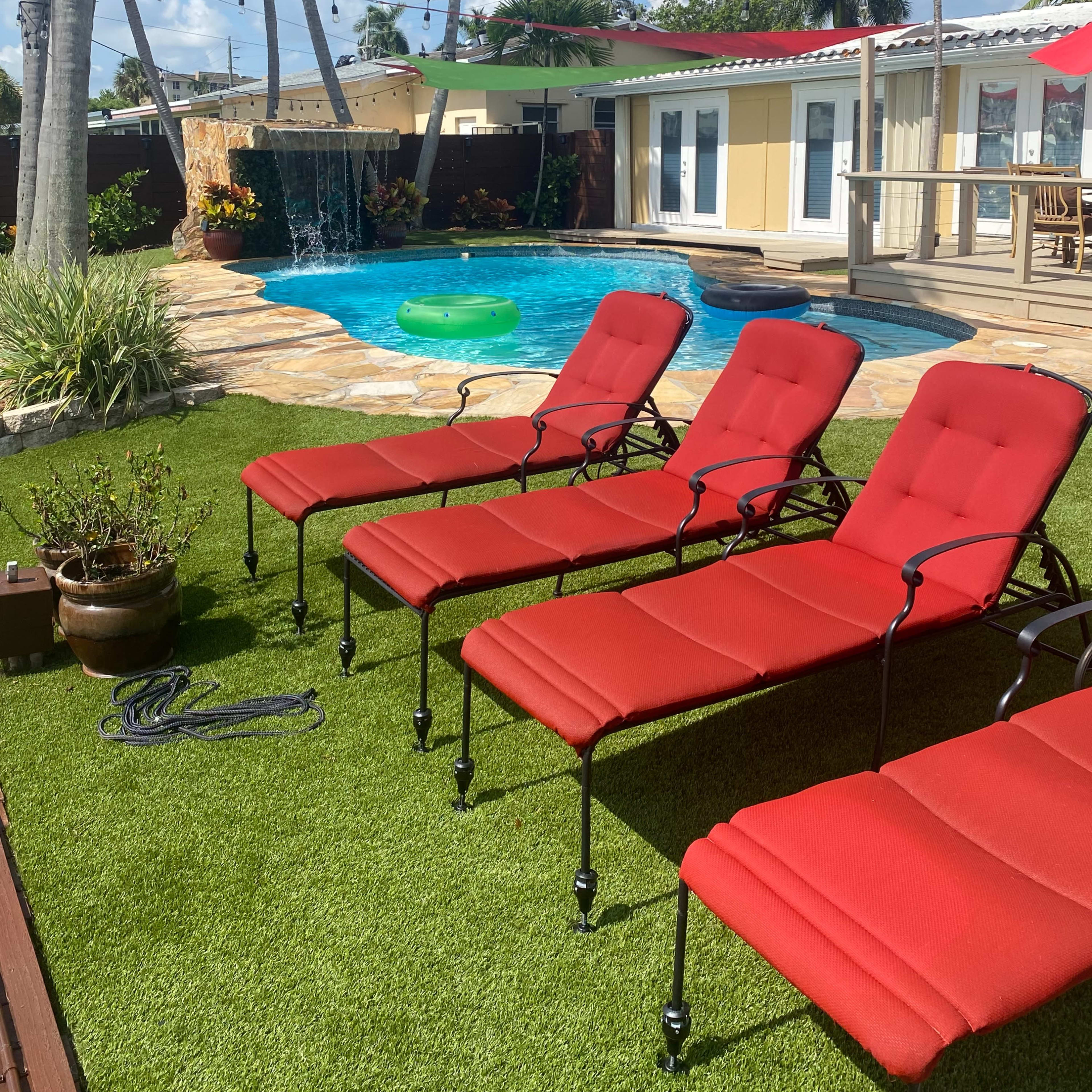 A sunny backyard scene features four red lounge chairs arranged near a swimming pool with a small waterfall and greenery.