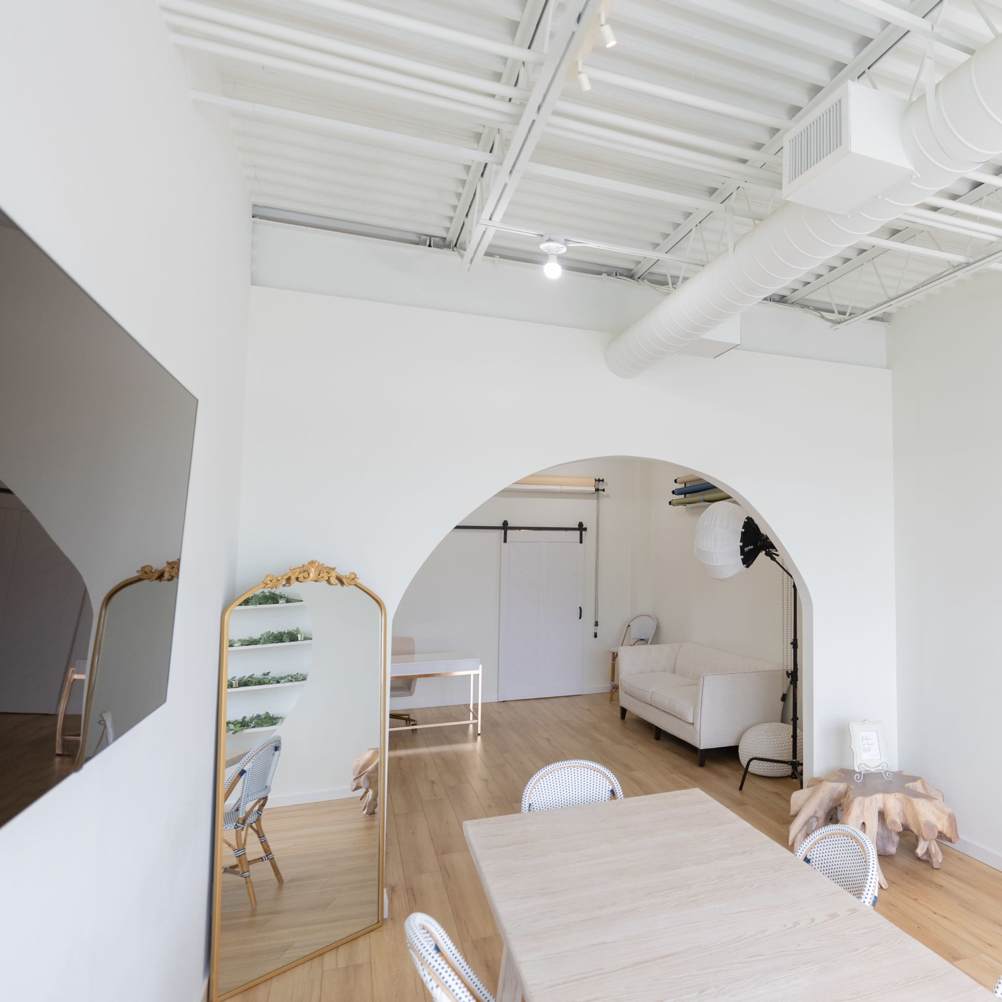 A bright, modern studio space featuring an arched wall with shelves adorned with greenery, a wooden dining table, and a large mirror.