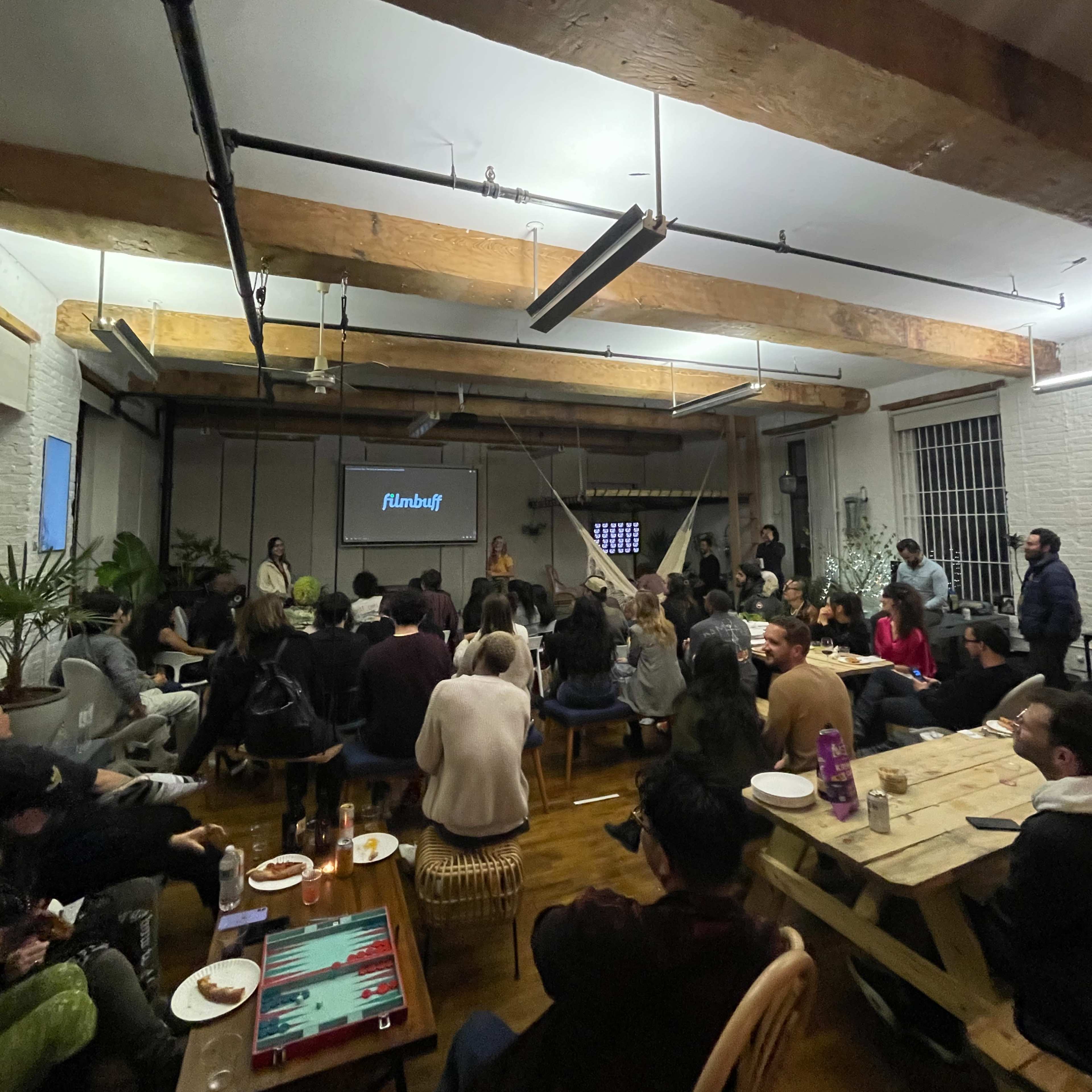 A group of people is seated in a spacious room with wooden beams, watching a presentation on a screen while food and drinks are placed on tables.