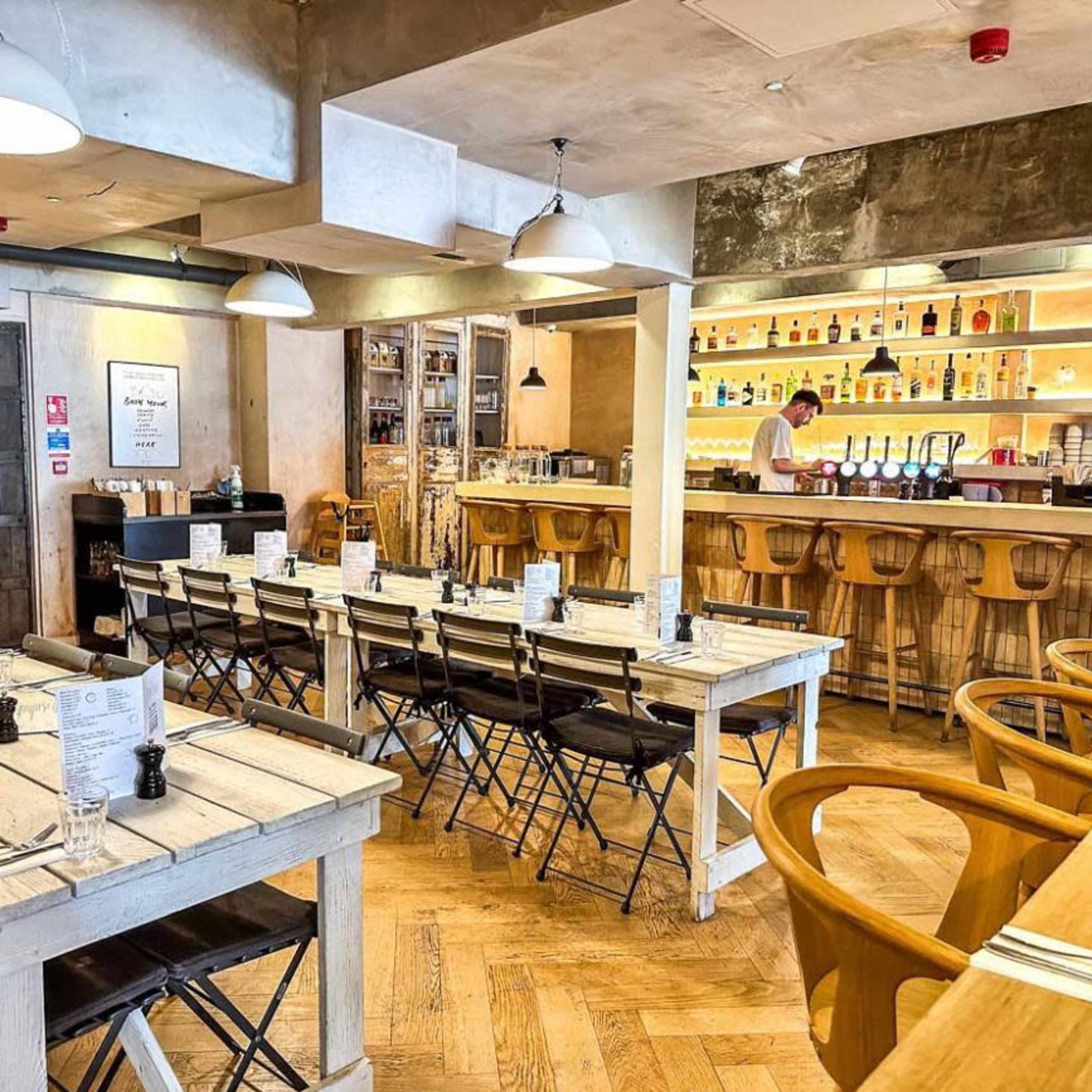 A modern restaurant interior with wooden tables, black chairs, and a bar featuring shelves of bottles in the background.