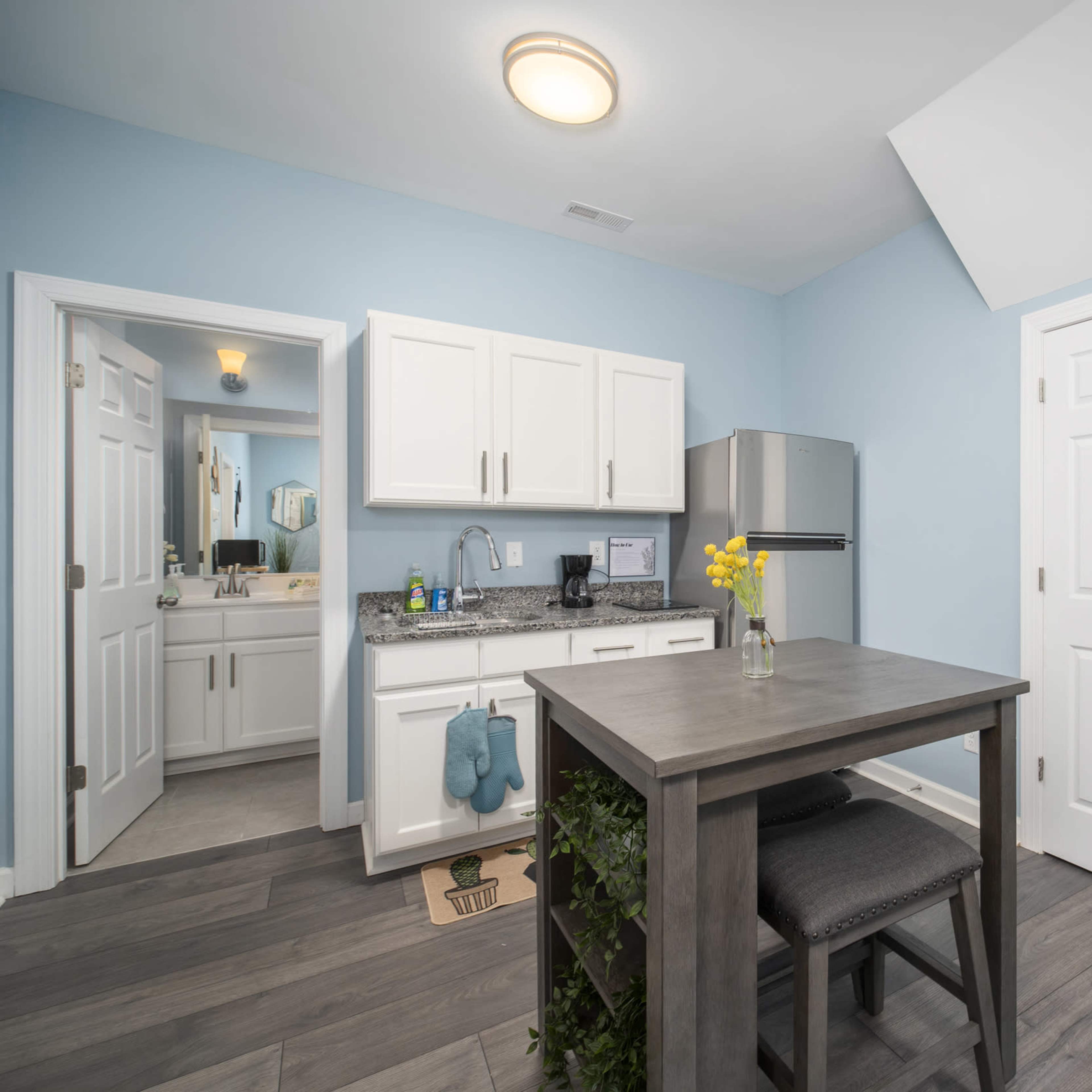 A small kitchen area with light blue walls, a wooden table with two chairs, a refrigerator, and a doorway leading to another room.