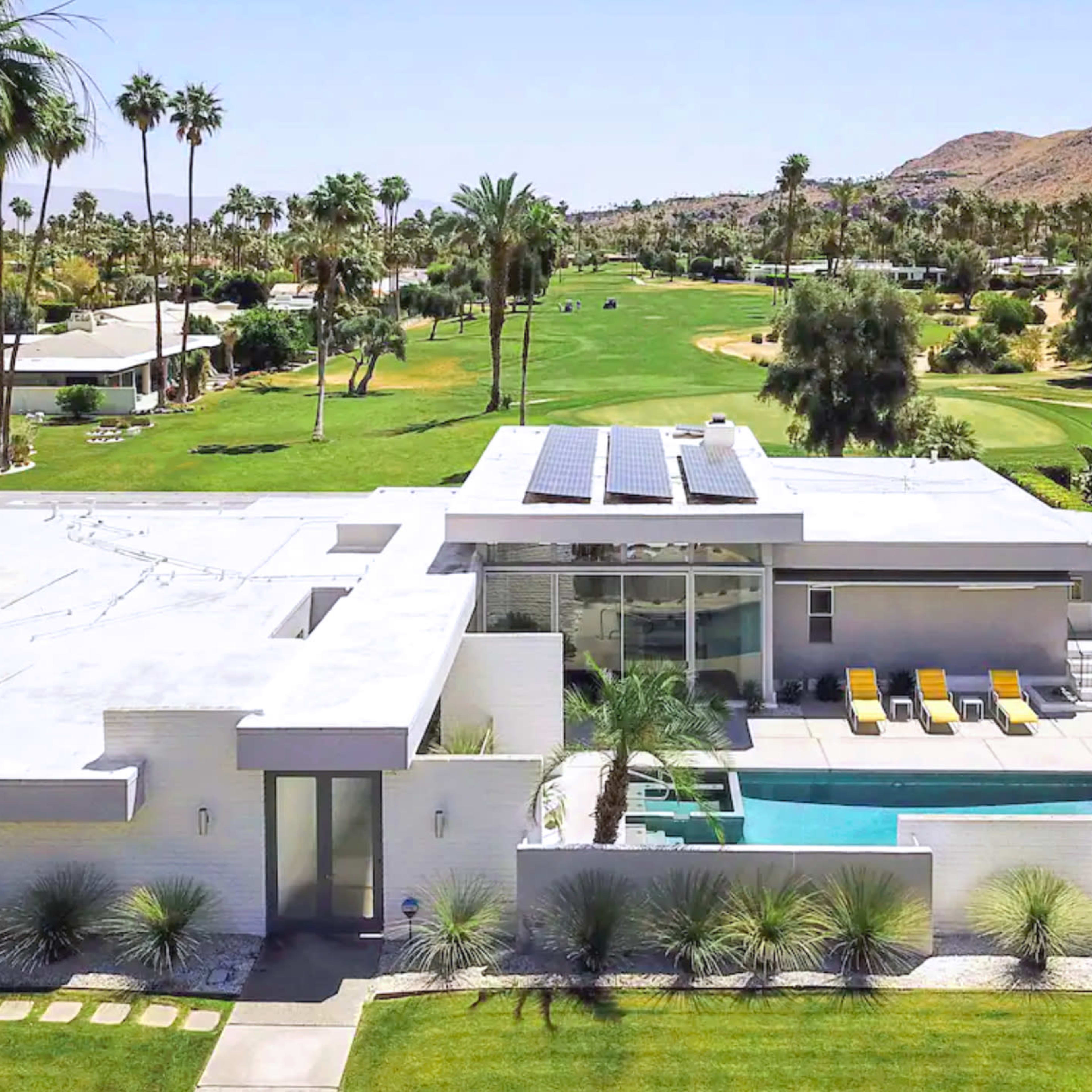 A modern single-story house with a swimming pool, solar panels on the roof, and a golf course in the background, surrounded by palm trees and landscaping.