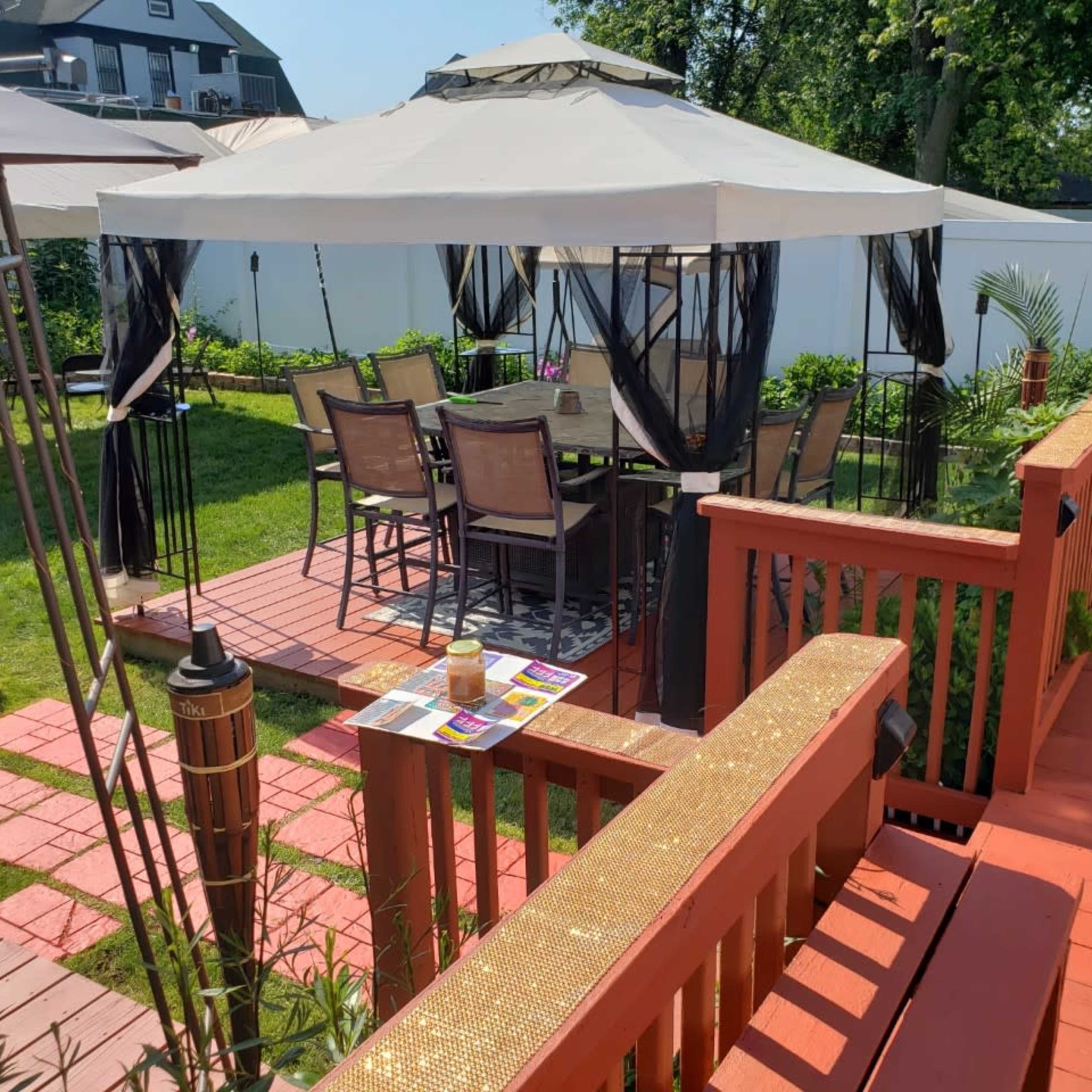 A backyard with a gazebo over a dining table surrounded by chairs, paved walkways, and a landscaped lawn.