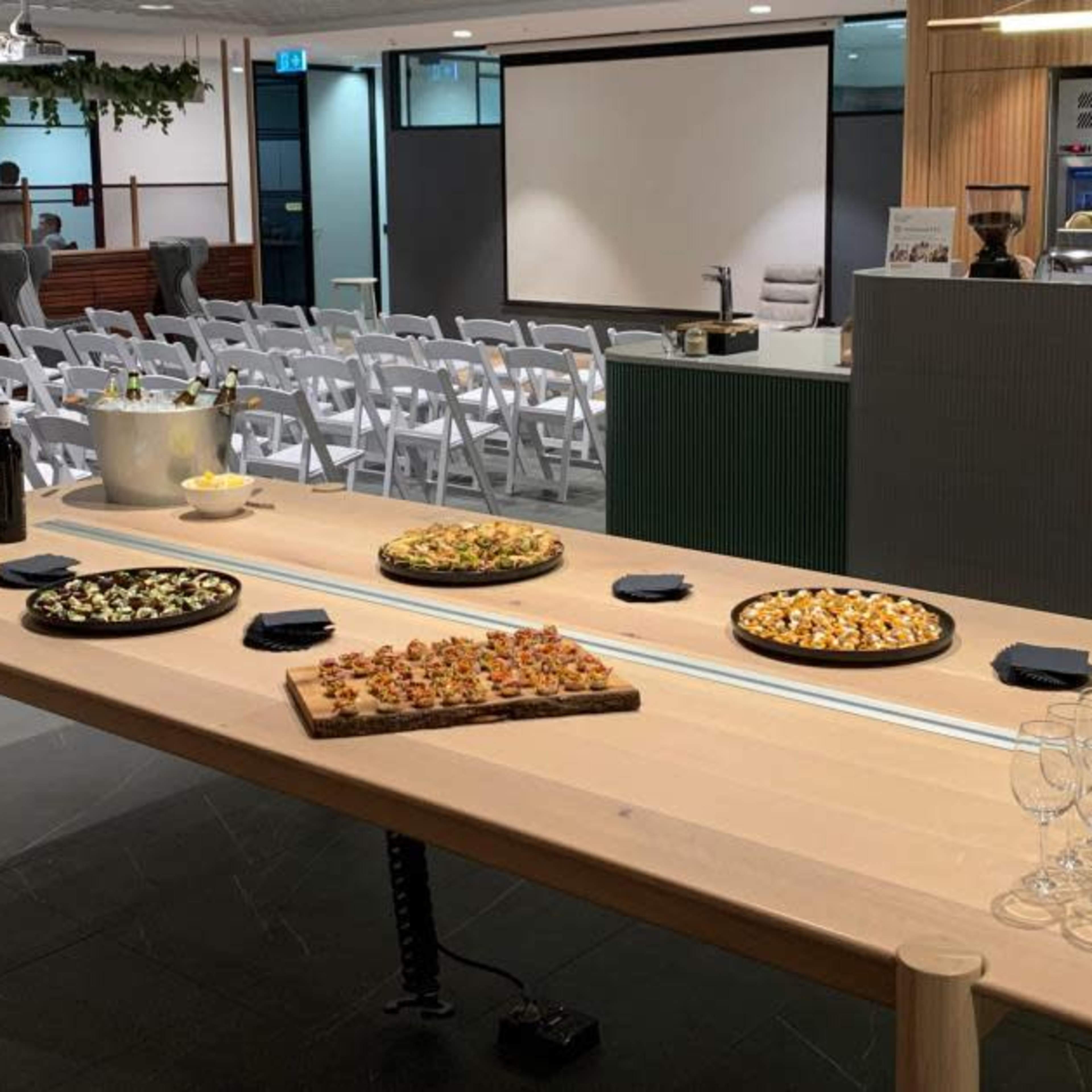 A long wooden table is set with various platters of food and drinks in a spacious room with chairs arranged for an event.