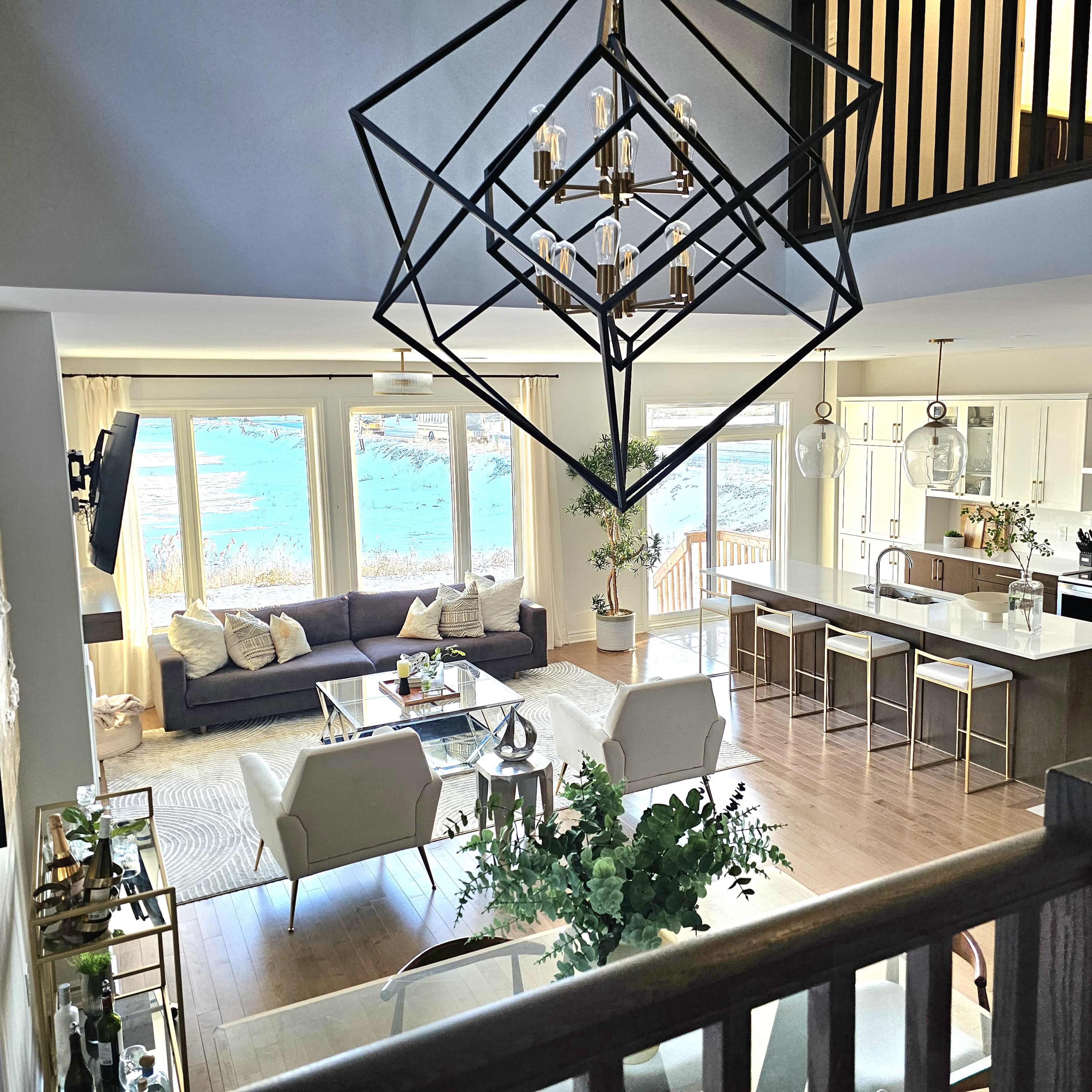 A modern living room with a view of water, featuring a gray sofa, white chairs, a coffee table, and a large chandelier overhead.