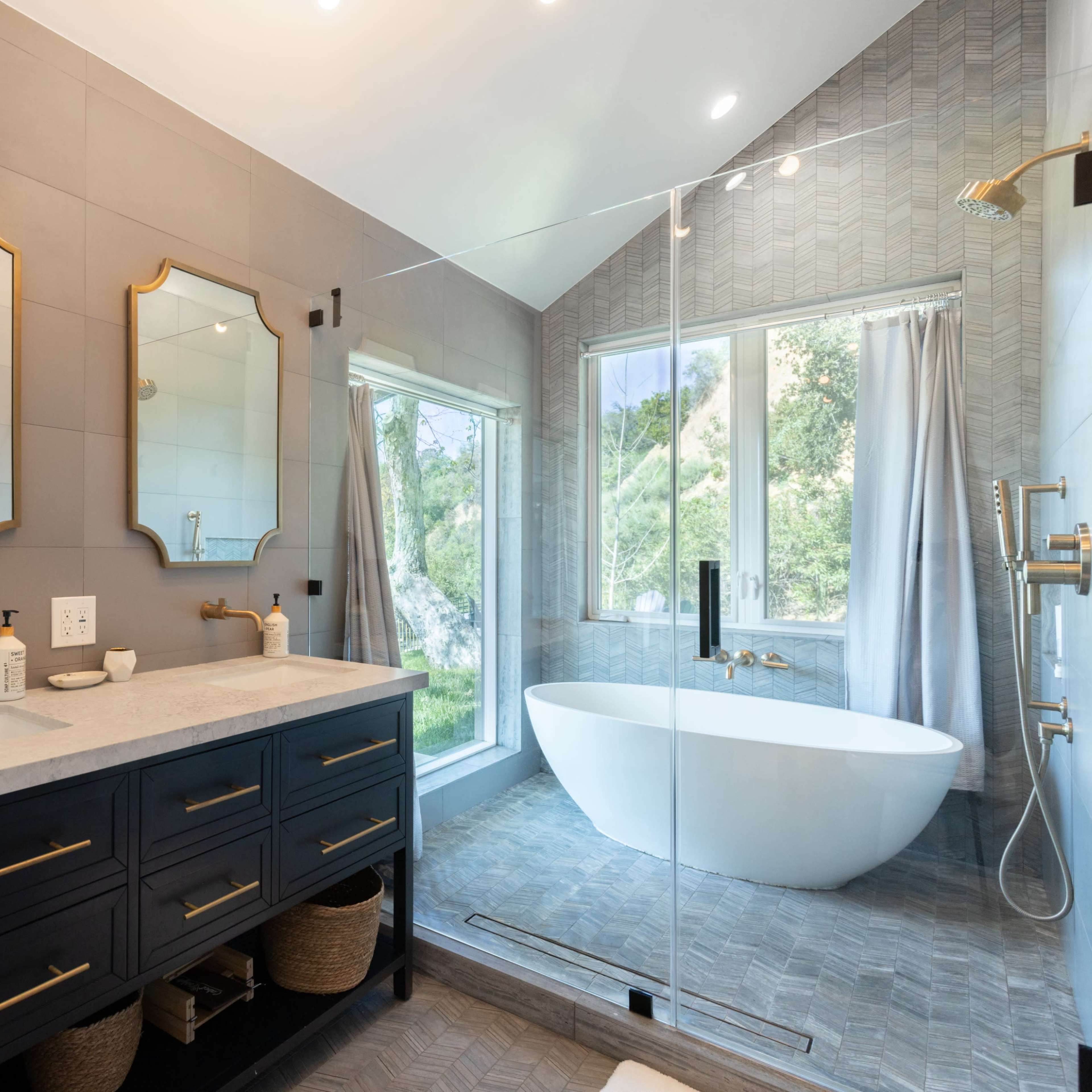 A modern bathroom features a double sink vanity with mirrors, a freestanding bathtub, and large windows overlooking greenery.