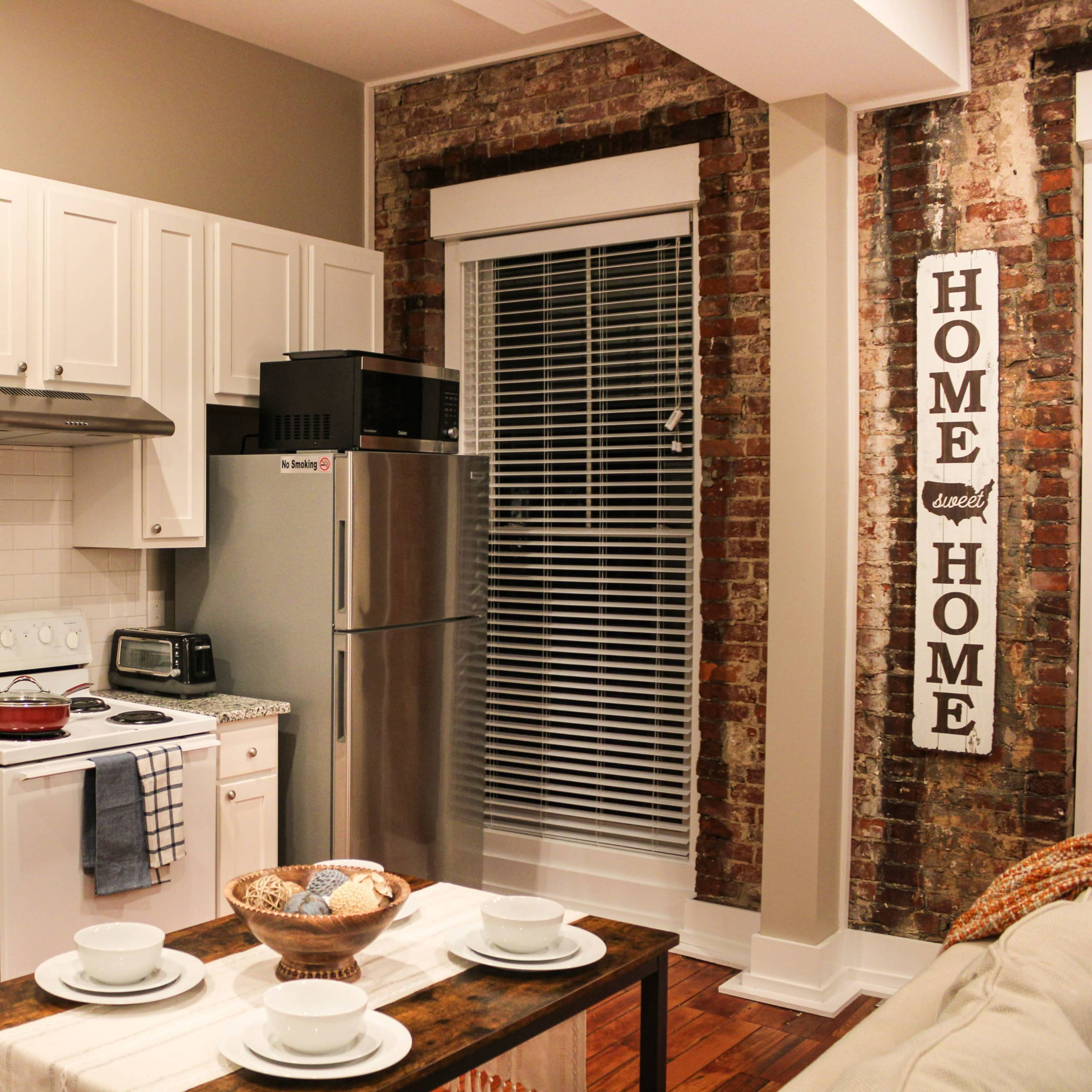 A modern kitchen features white cabinetry, a stainless steel refrigerator, and exposed brick walls.