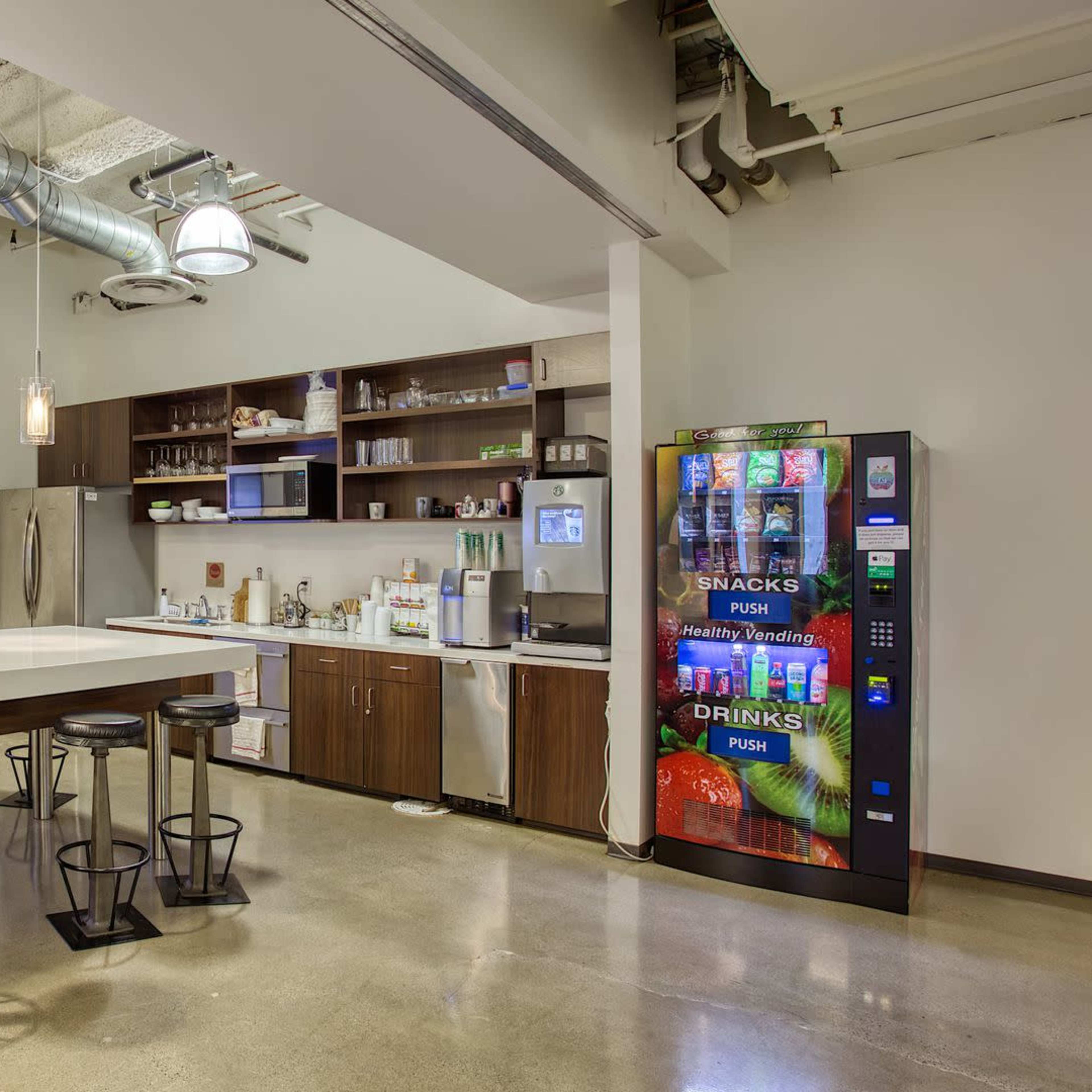 A modern break room with a large white table, bar stools, kitchen appliances, and a vending machine.