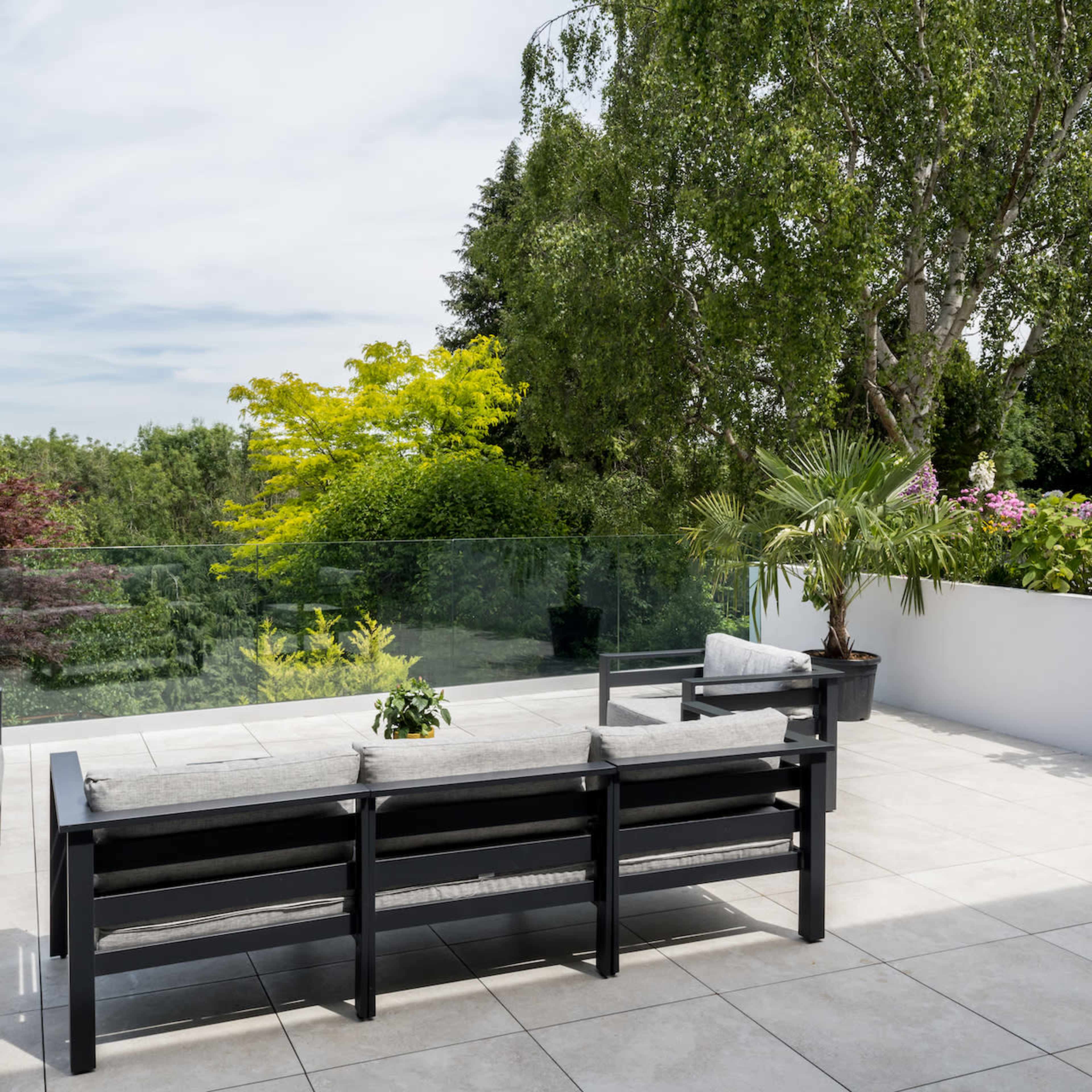A modern outdoor patio features a black sectional sofa with cushions, surrounded by greenery and flowering plants.