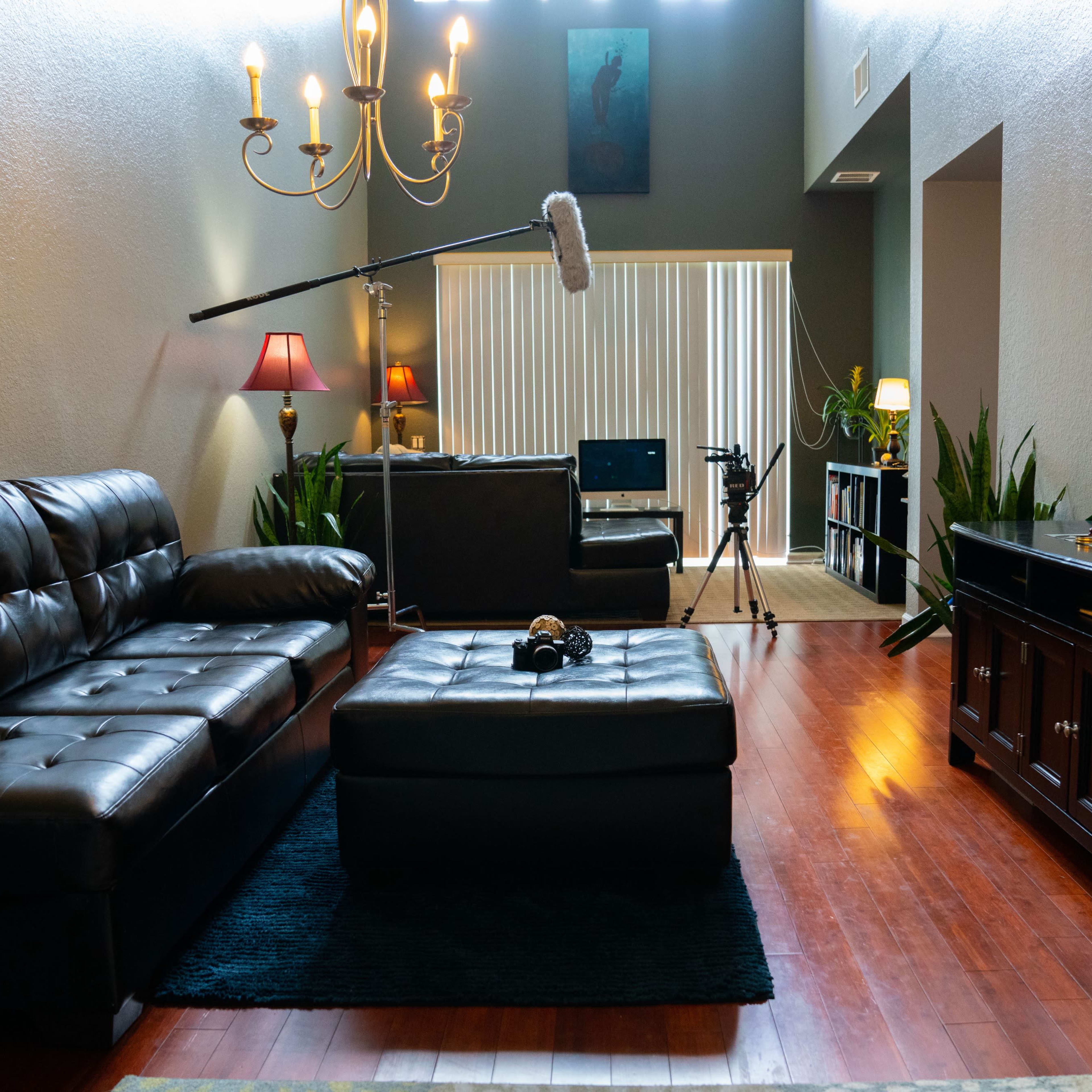 A living room with dark leather sofas, a coffee table in the center, a chandelier overhead, and a camera setup in the background.